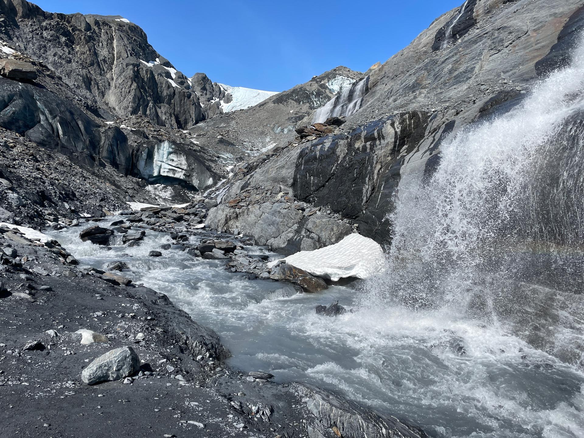 Powerful waterfall cascades into a rocky stream with a glacier visible in the background.