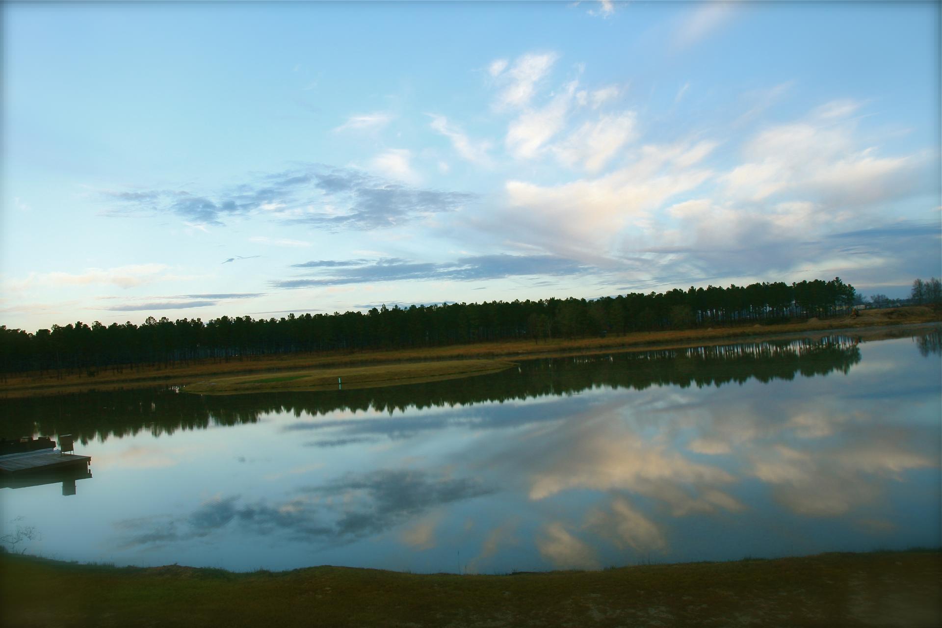 Calm lake reflecting cloudy sky and distant evergreen trees at dusk.