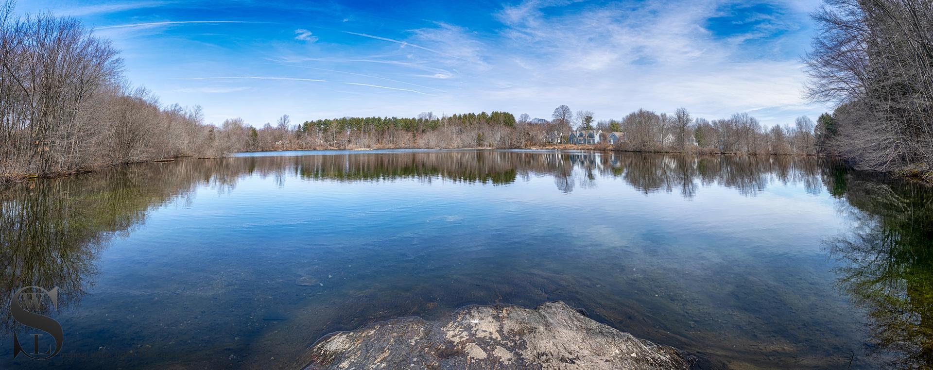 Panoramic view of a calm lake reflecting bare trees under a blue sky.