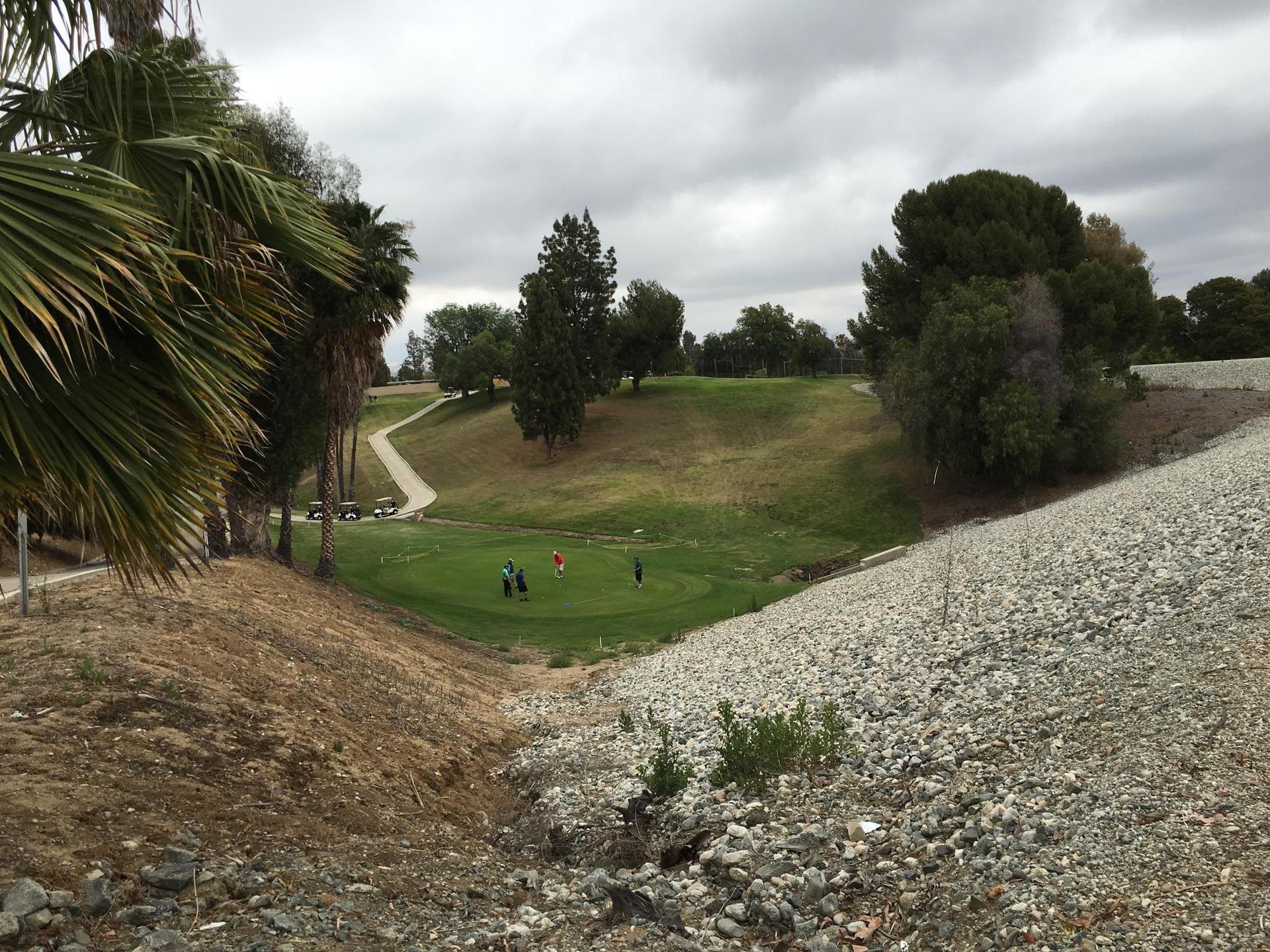 Overcast view of golfers on a green course, surrounded by trees and rocky slopes.