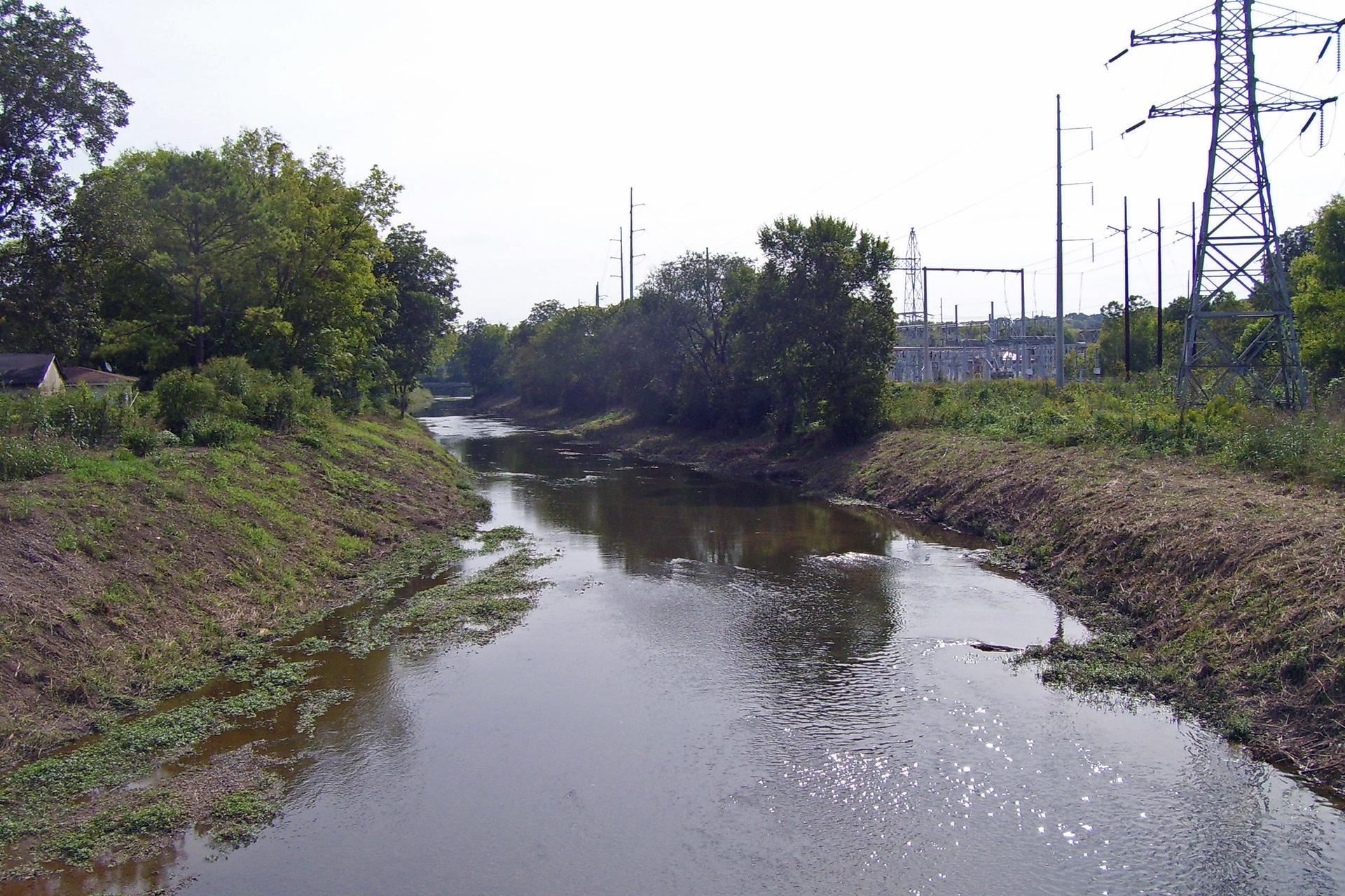 A winding river with green banks, trees, and power lines in the distance.