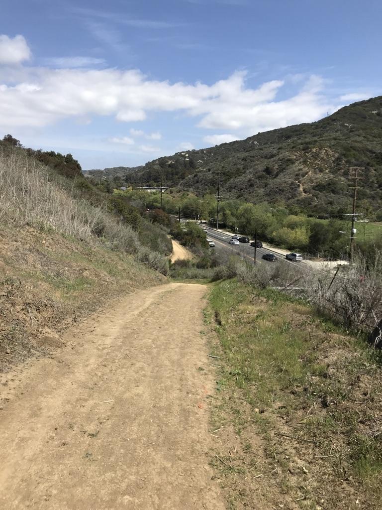 A dirt path leads down to a road with cars, surrounded by green hills.
