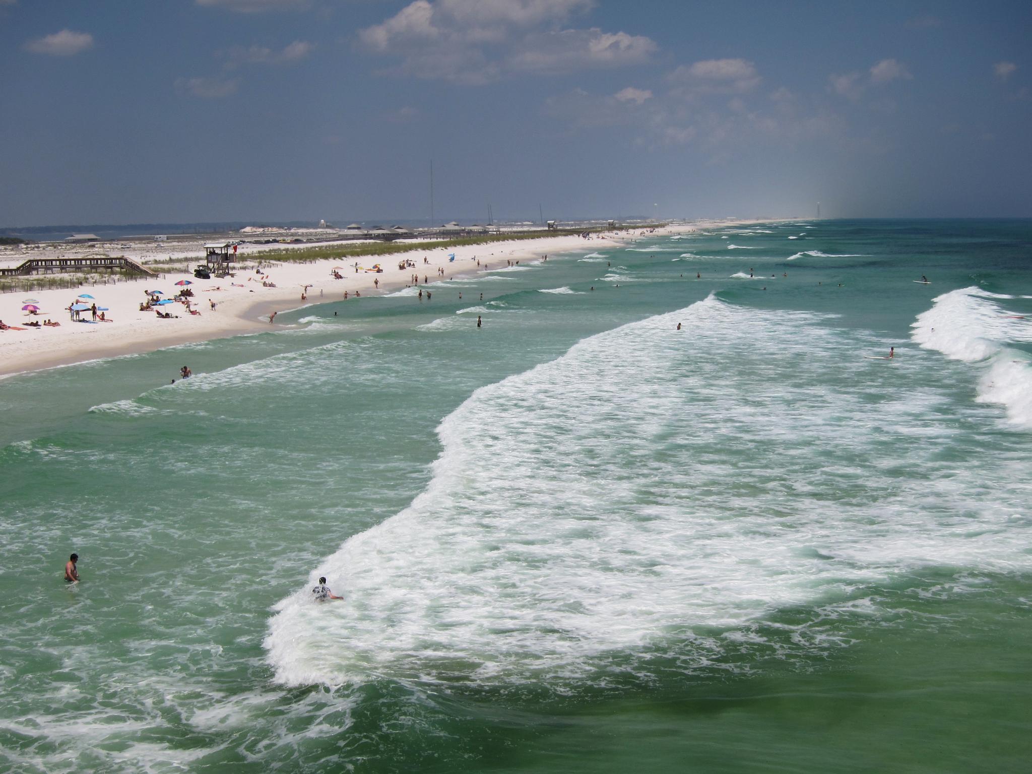 Sunny beach with white sand, turquoise water, and people swimming and sunbathing.
