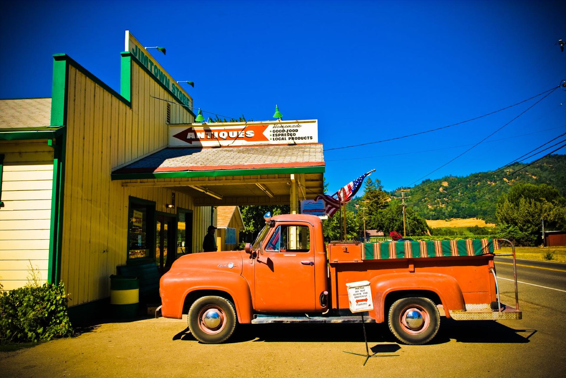An orange vintage pickup truck parked outside an antique store on a sunny day.