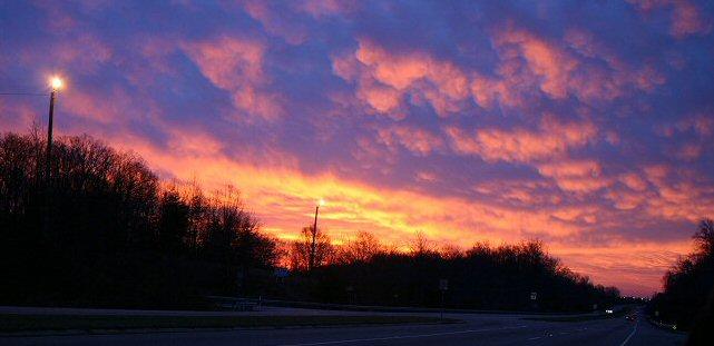 A stunning sunset with fiery clouds above a dark road and trees.