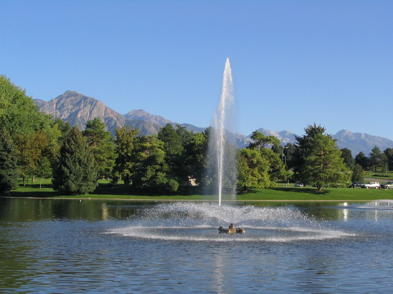 A tall fountain sprays water in a lake with green trees and mountains under a clear sky. - Sugar House Park Loop