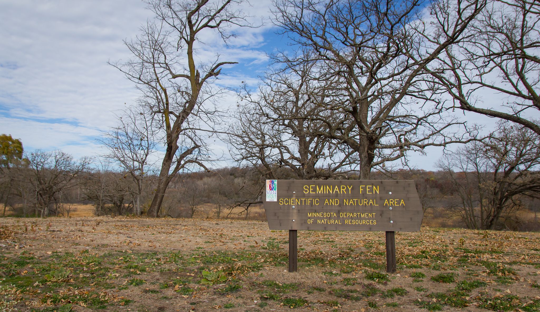 Wooden sign in a dry, grassy field with bare trees under a cloudy sky. - Seminary Fen SNA – Bluff Creek Drive trail access loop from Bavaria Rd