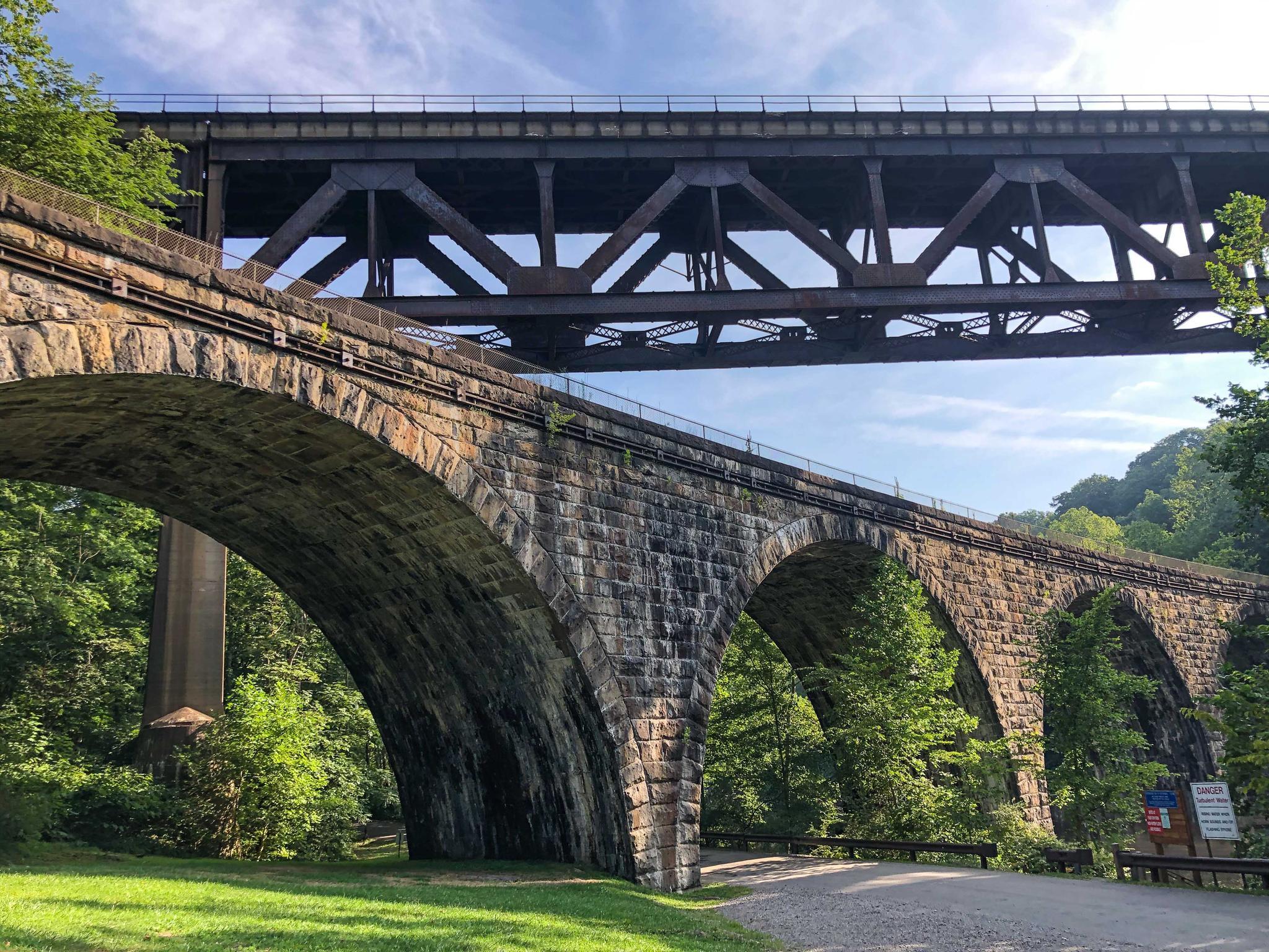 Two bridges, one stone arch and one metal truss, span a green valley.