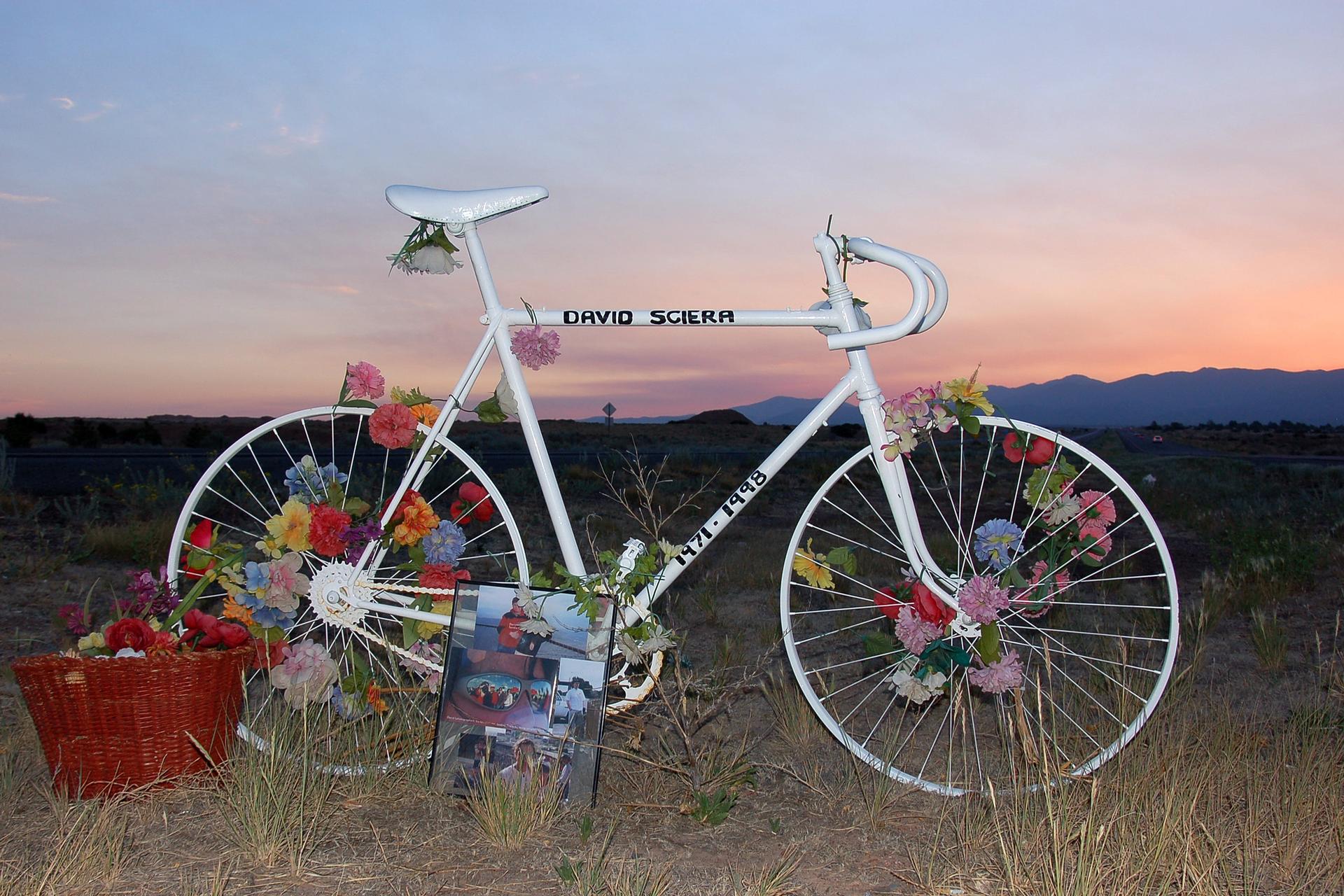 White ghost bike memorial with colorful flowers, basket, and picture at sunset.