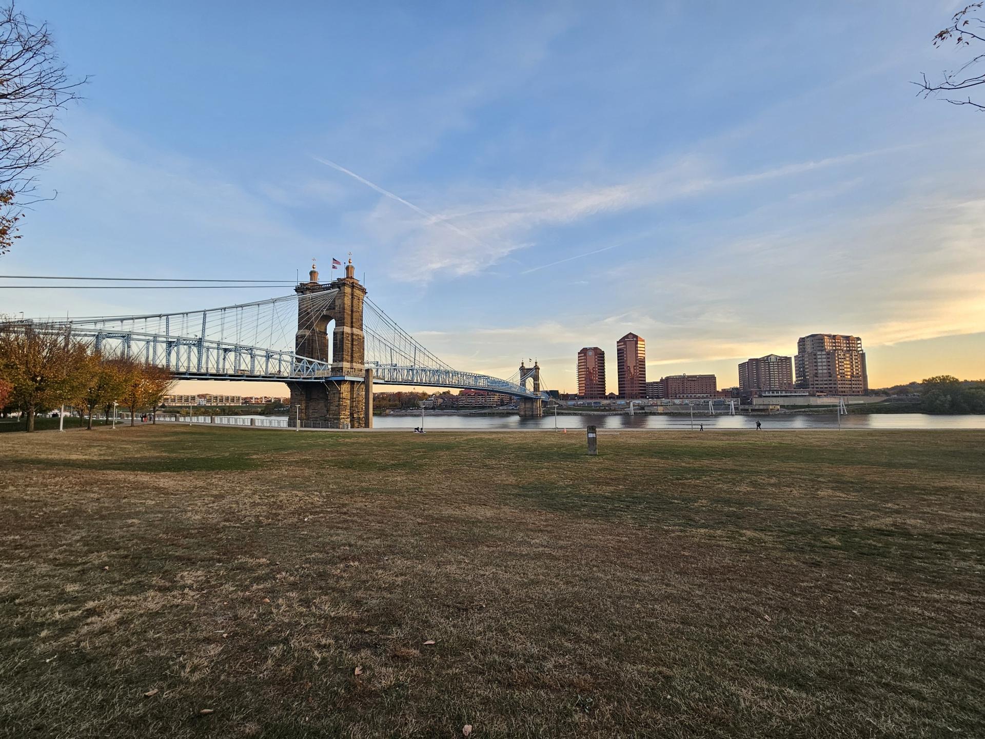 Suspension bridge and city skyline across river from a grassy park.