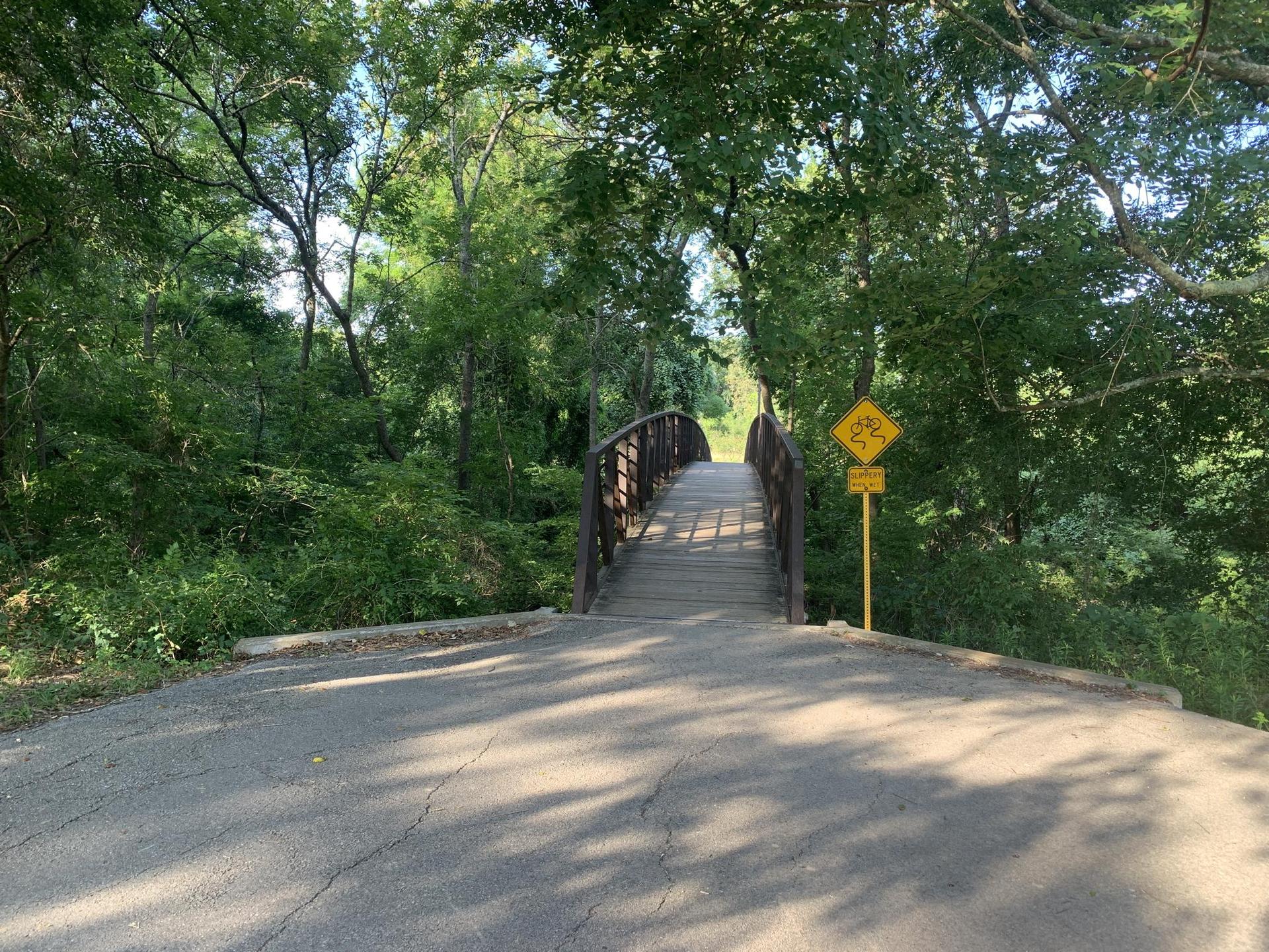 Paved road leading to a wooden bridge surrounded by trees.