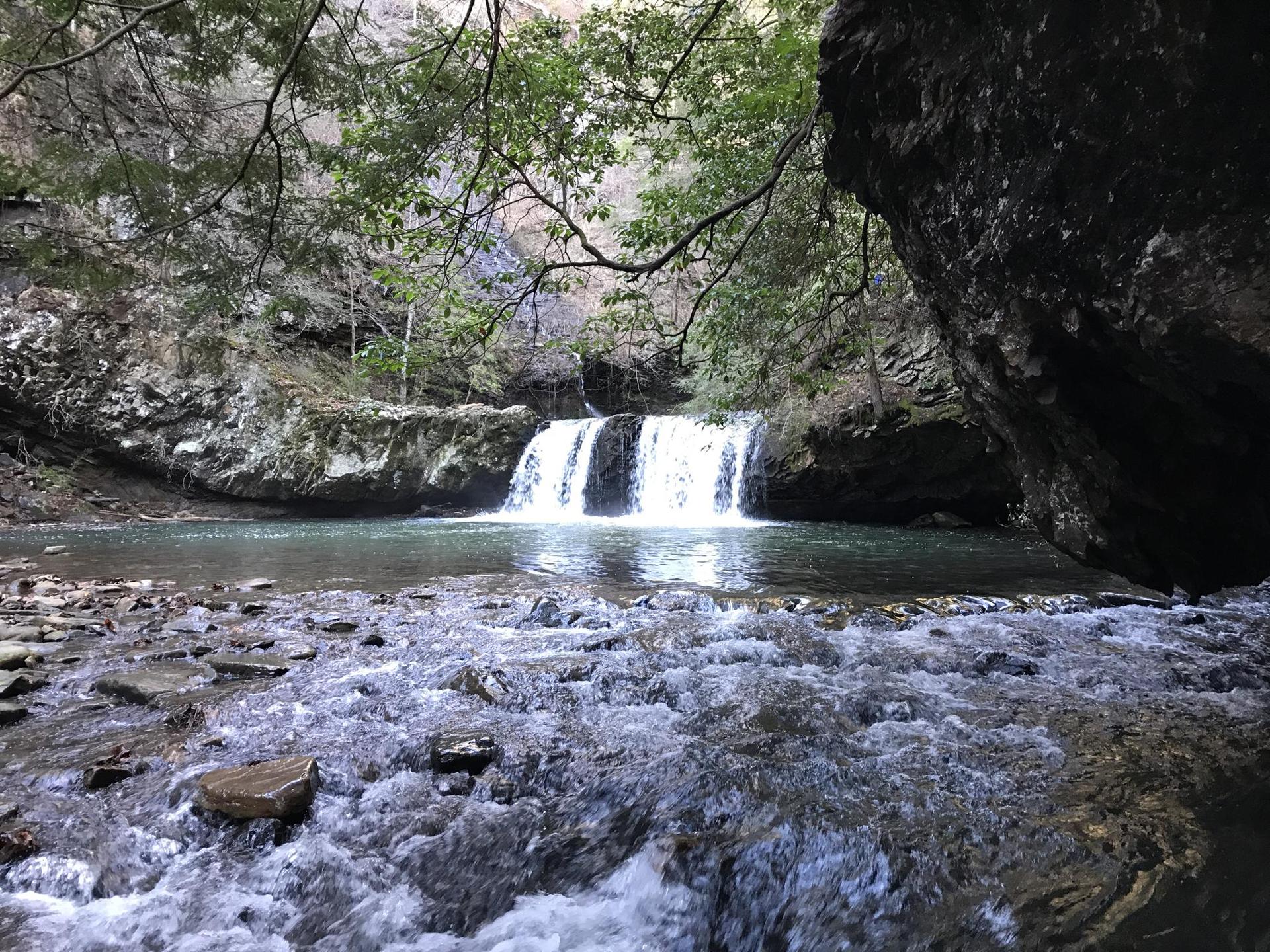 A multi-tiered waterfall flows into a rocky river under dappled light.