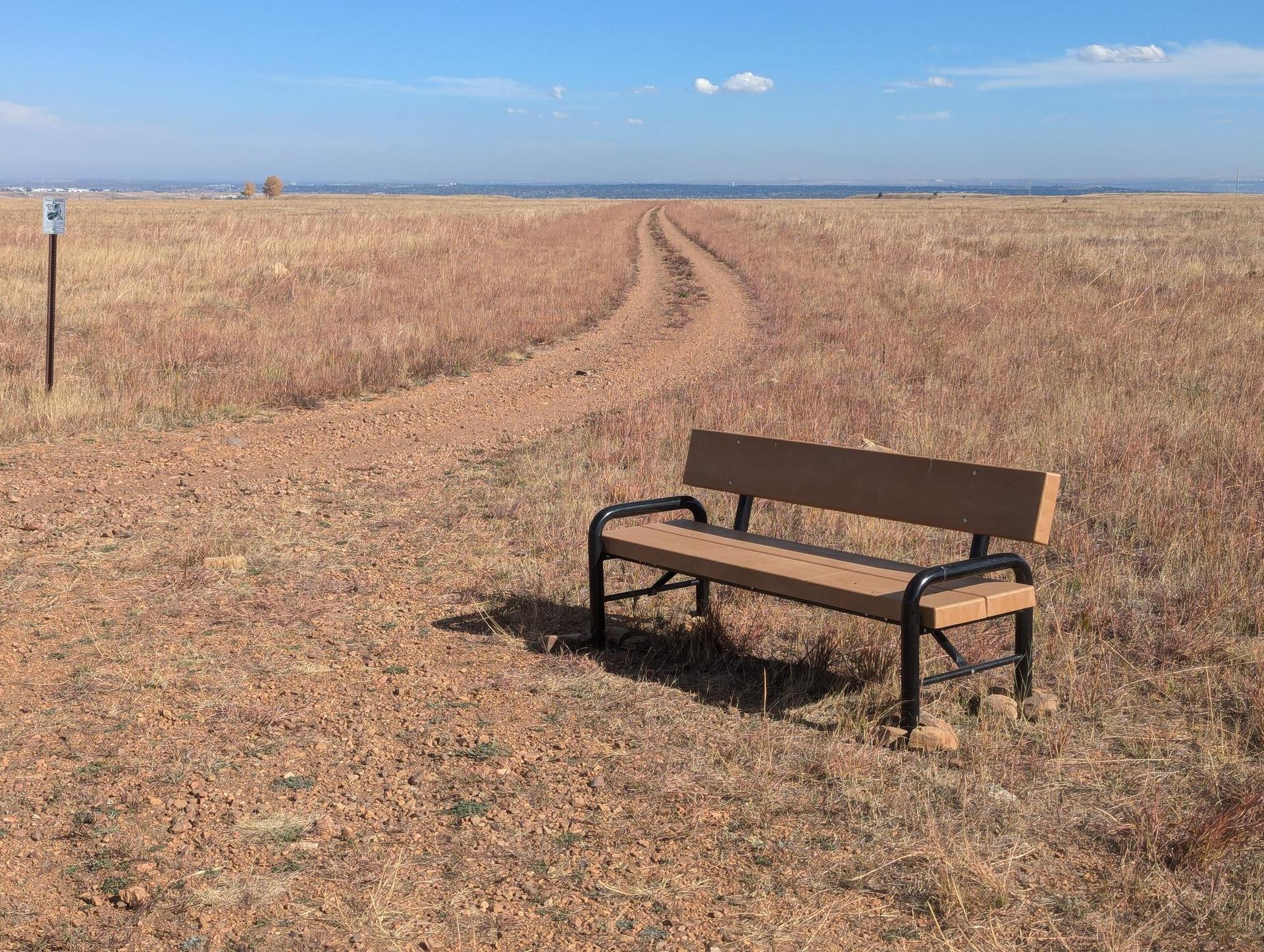 A wooden bench sits beside a winding dirt path through dry golden fields.