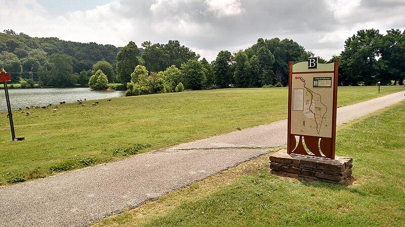An information sign next to a paved path by a lake and grassy field.