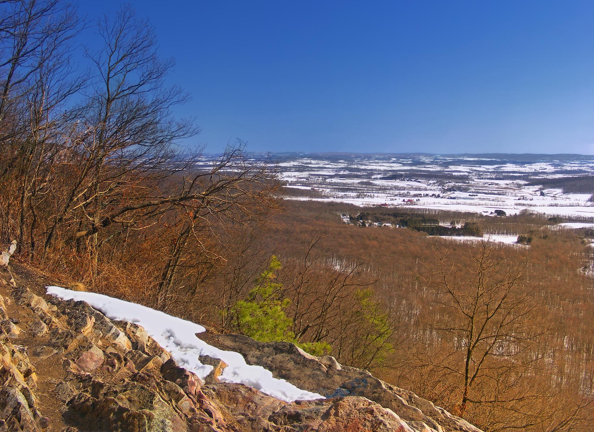 Rocky foreground with snow overlooks a vast winter landscape under a clear blue sky.