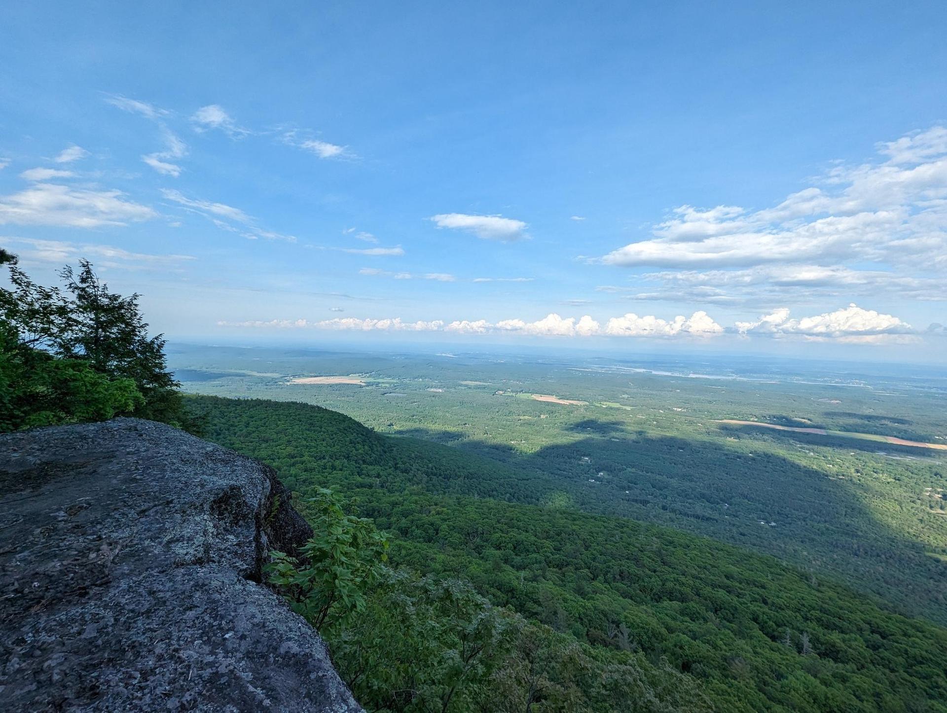 A vast green valley viewed from a rocky cliff under a blue sky with clouds.
