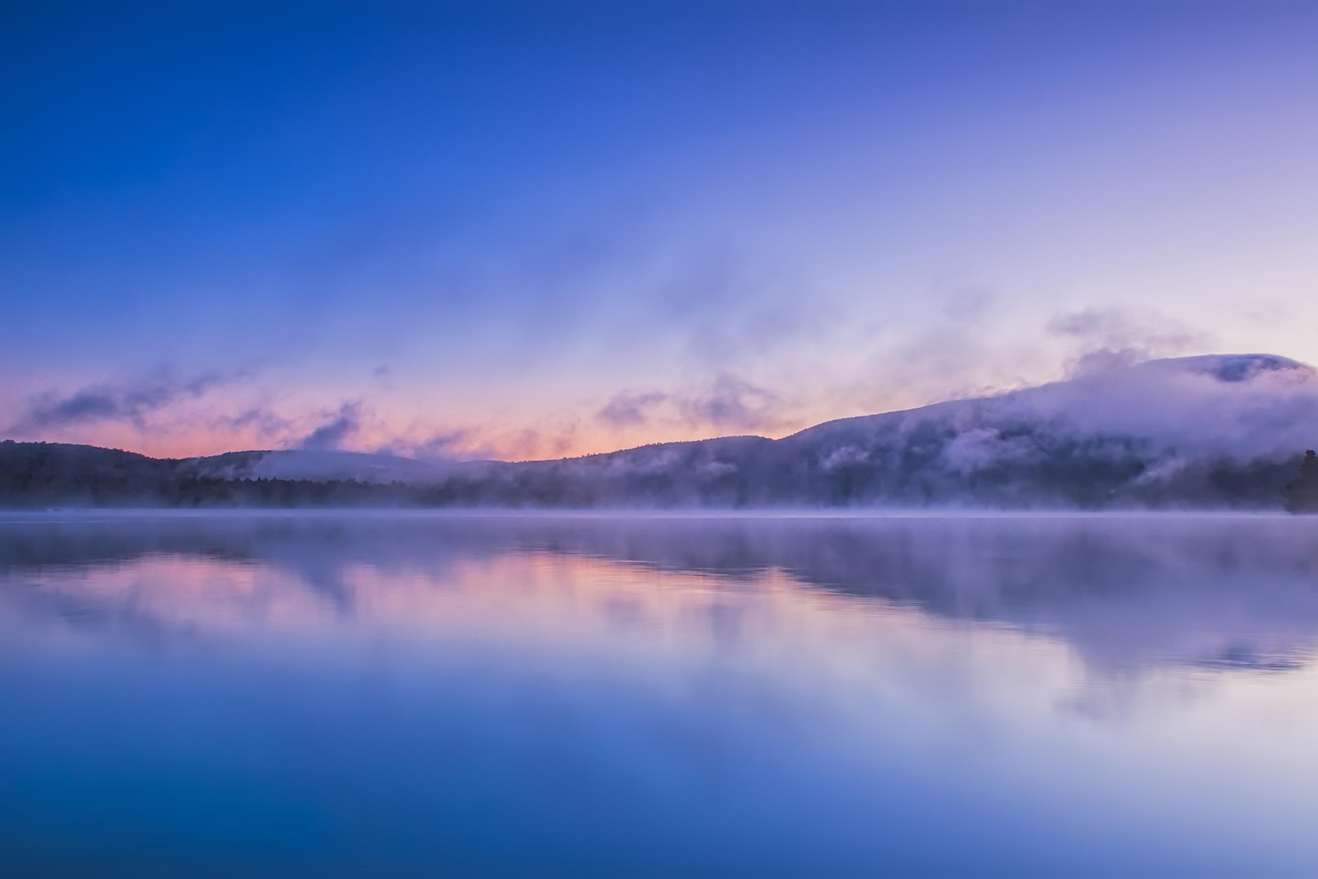 Misty lake reflecting colorful twilight sky with fog-covered mountains in the background.