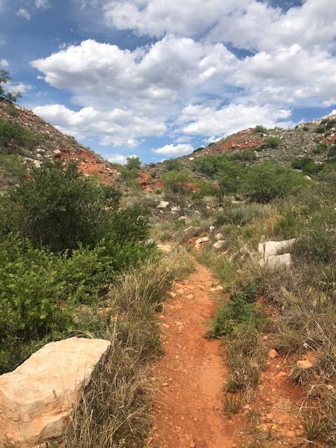 A dirt path winds through rocky, green hills under a partly cloudy sky.