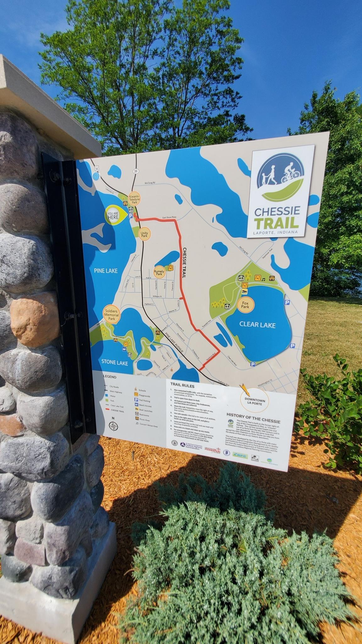 A large trail map sign stands next to a stone pillar under a bright blue sky.