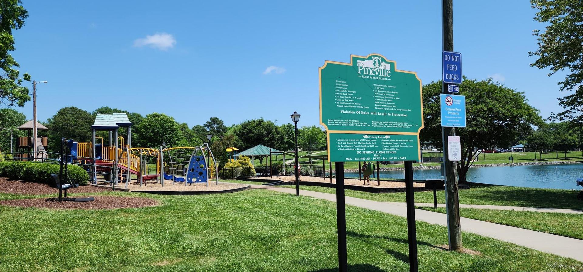 A sunny playground with a sign, green grass, and a pond.