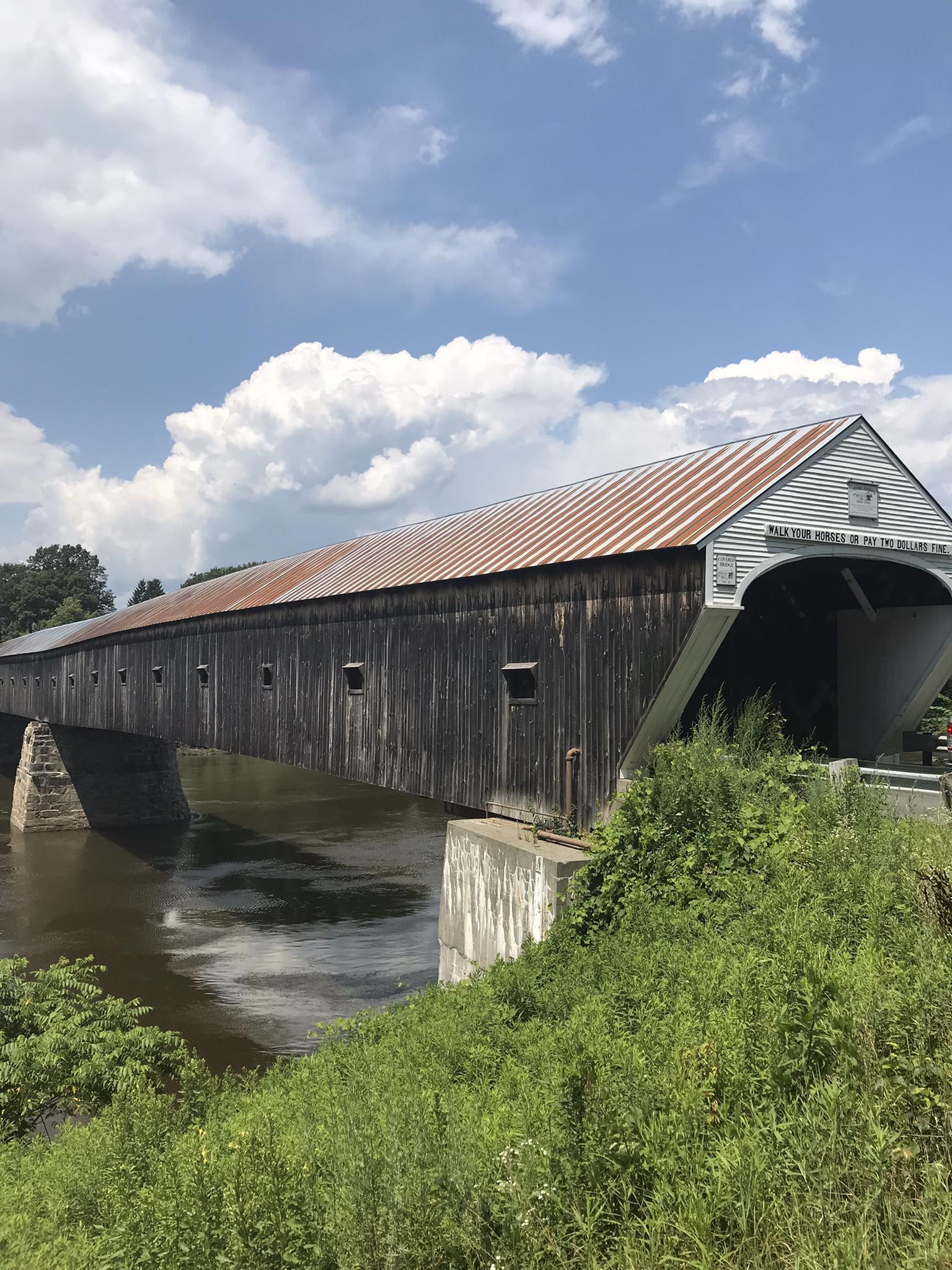 Wooden covered bridge crosses a river on a bright, cloudy day.