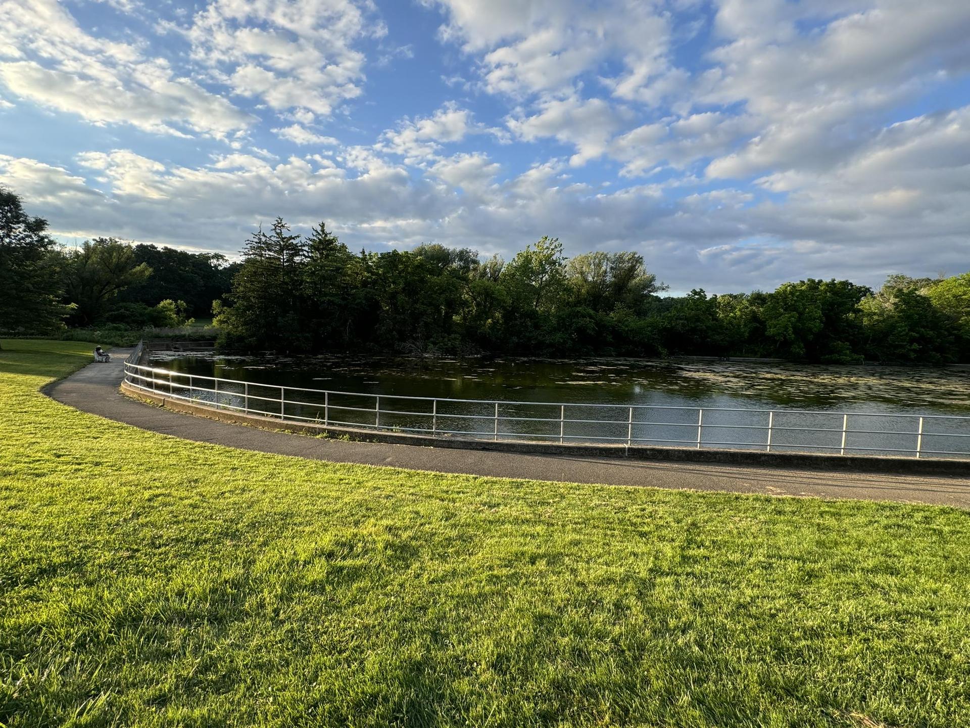 A lake with a railing, surrounded by green grass and trees, under a partly cloudy sky.