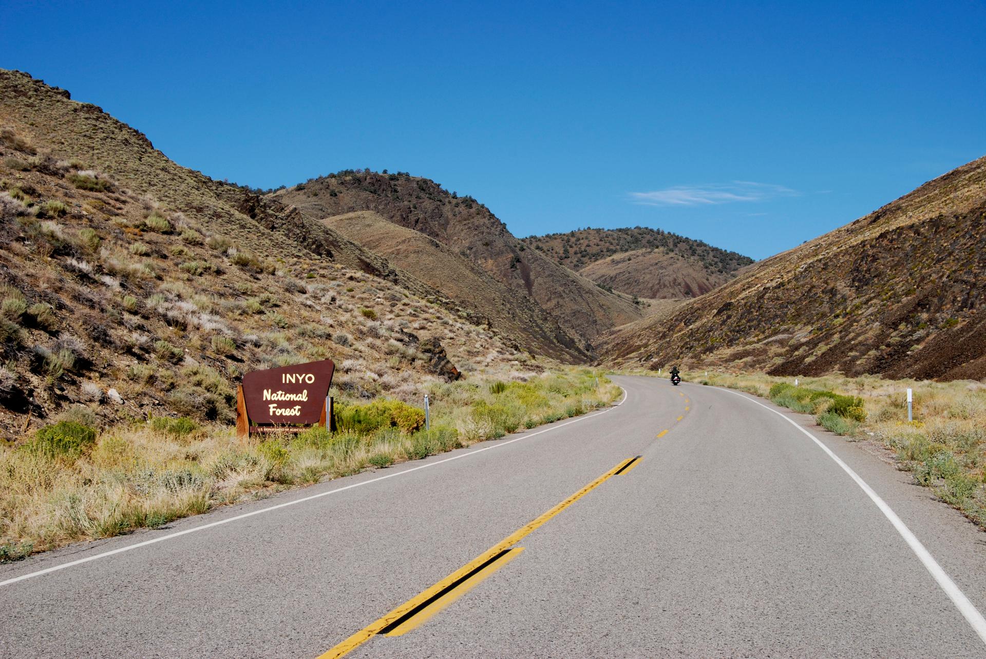 A road sign beside a winding road through arid mountains under a clear sky.