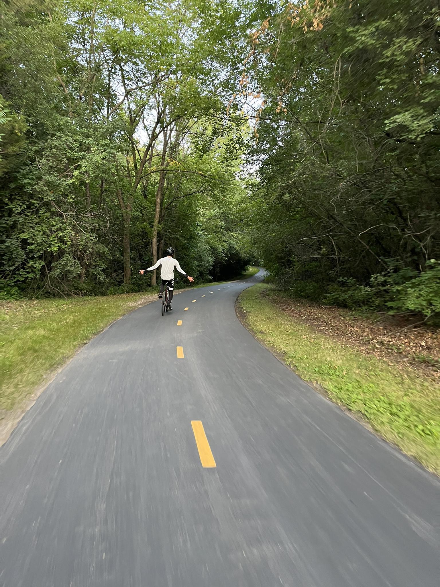 Person cycling with arms out on a paved path through a green forest.