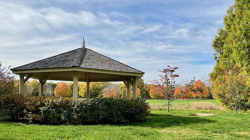 Gazebo surrounded by green bushes and autumn trees under a bright, cloudy sky.