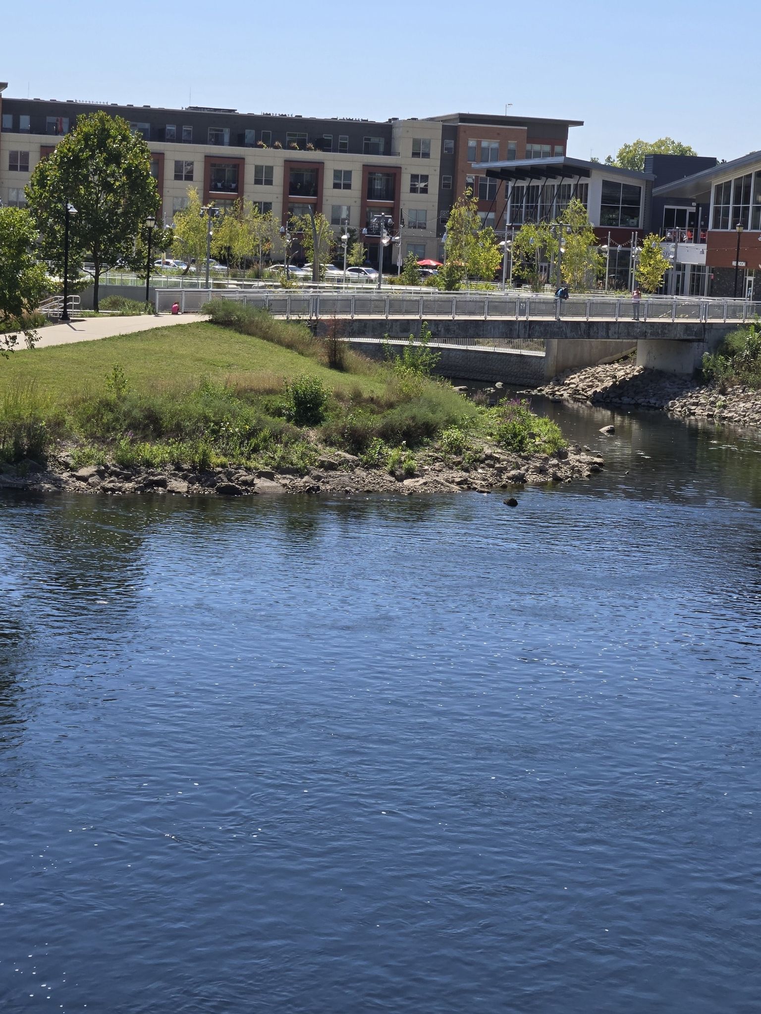 River flowing past a modern bridge, buildings, and green grassy banks. - River St. Joseph of the Miamis Trail