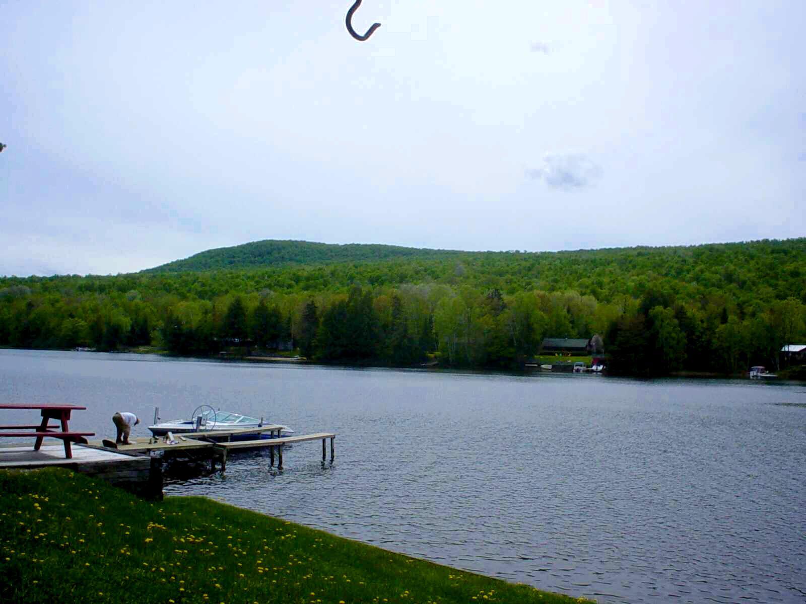A person works on a boat at a dock on a lake, with forested hills.