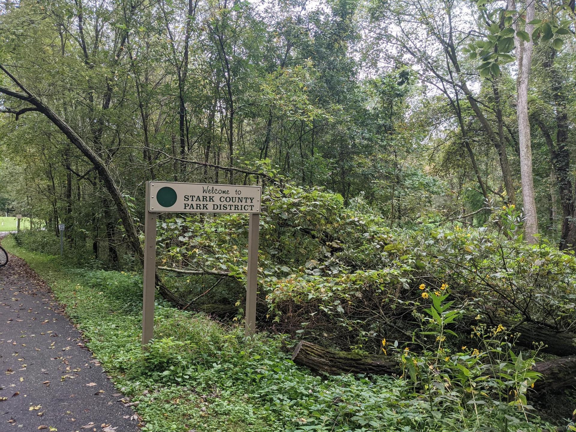 A wooden sign stands beside a paved path in a dense, green forest.