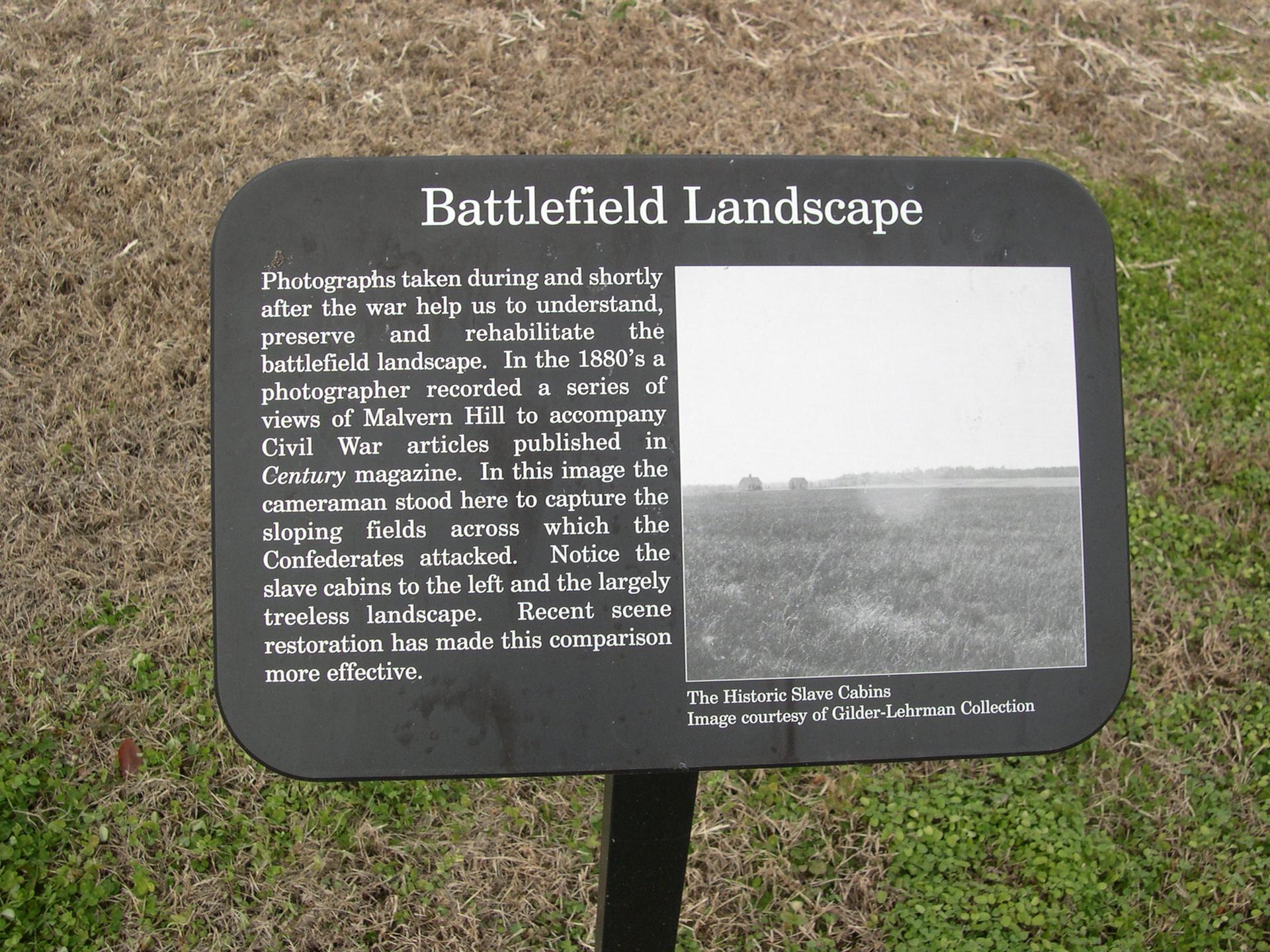 A black interpretive sign with text and a historical black and white photo on grassy ground.