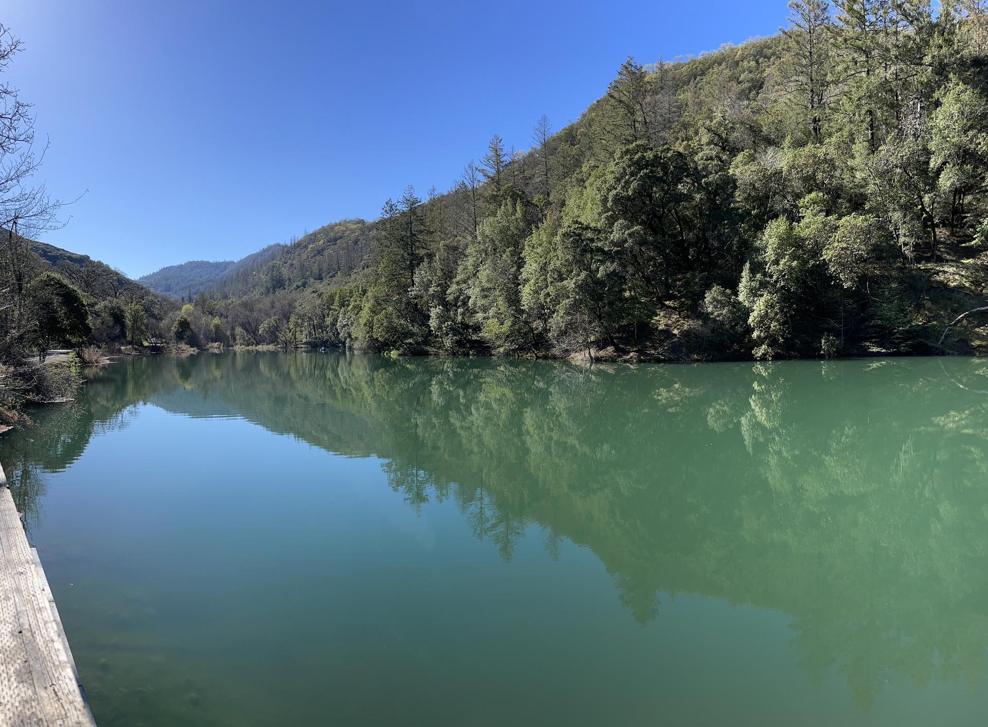 A calm green lake reflects forested hills under a bright blue sky.