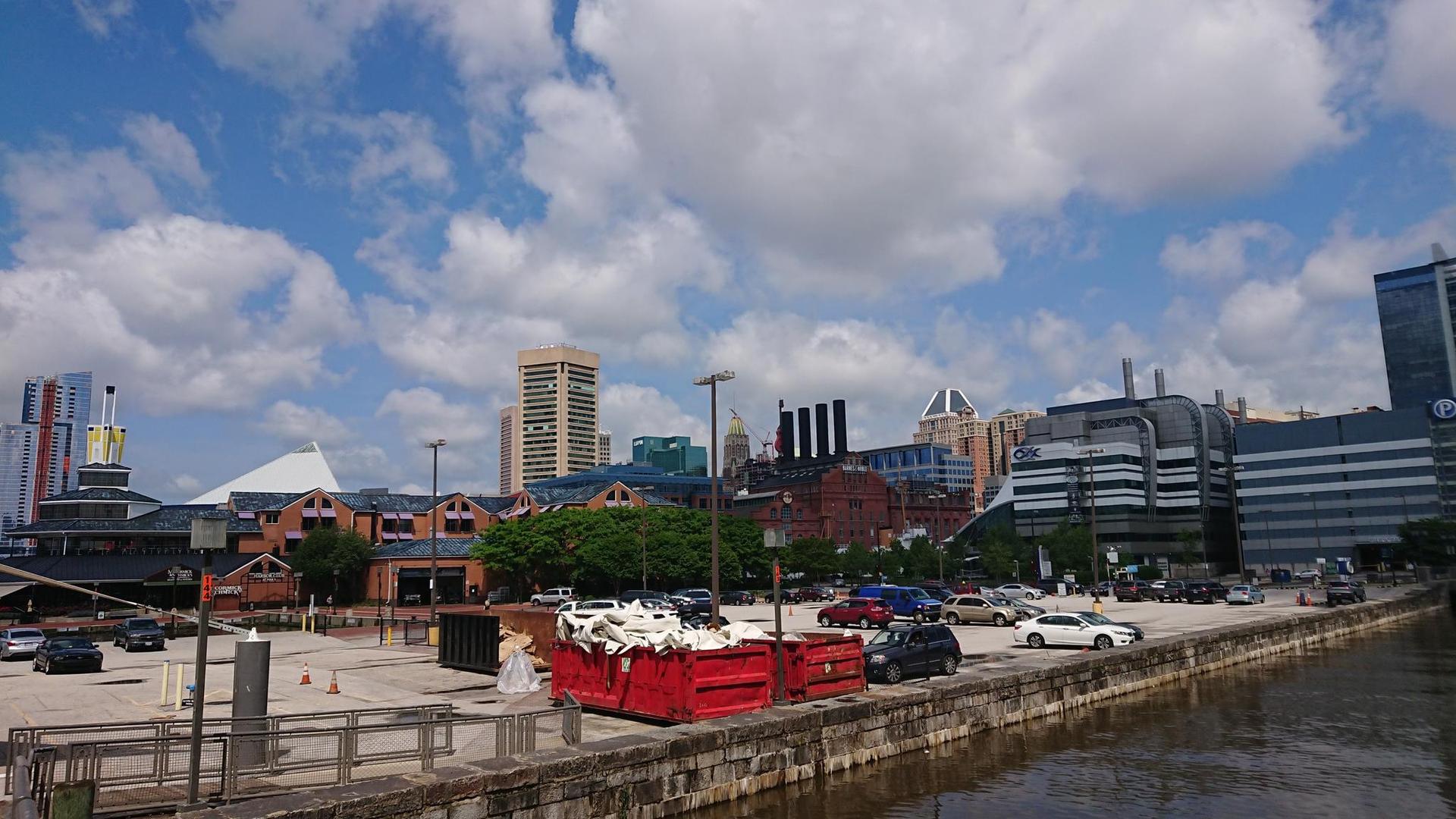 A red dumpster on a paved waterfront, with city buildings and a cloudy sky.