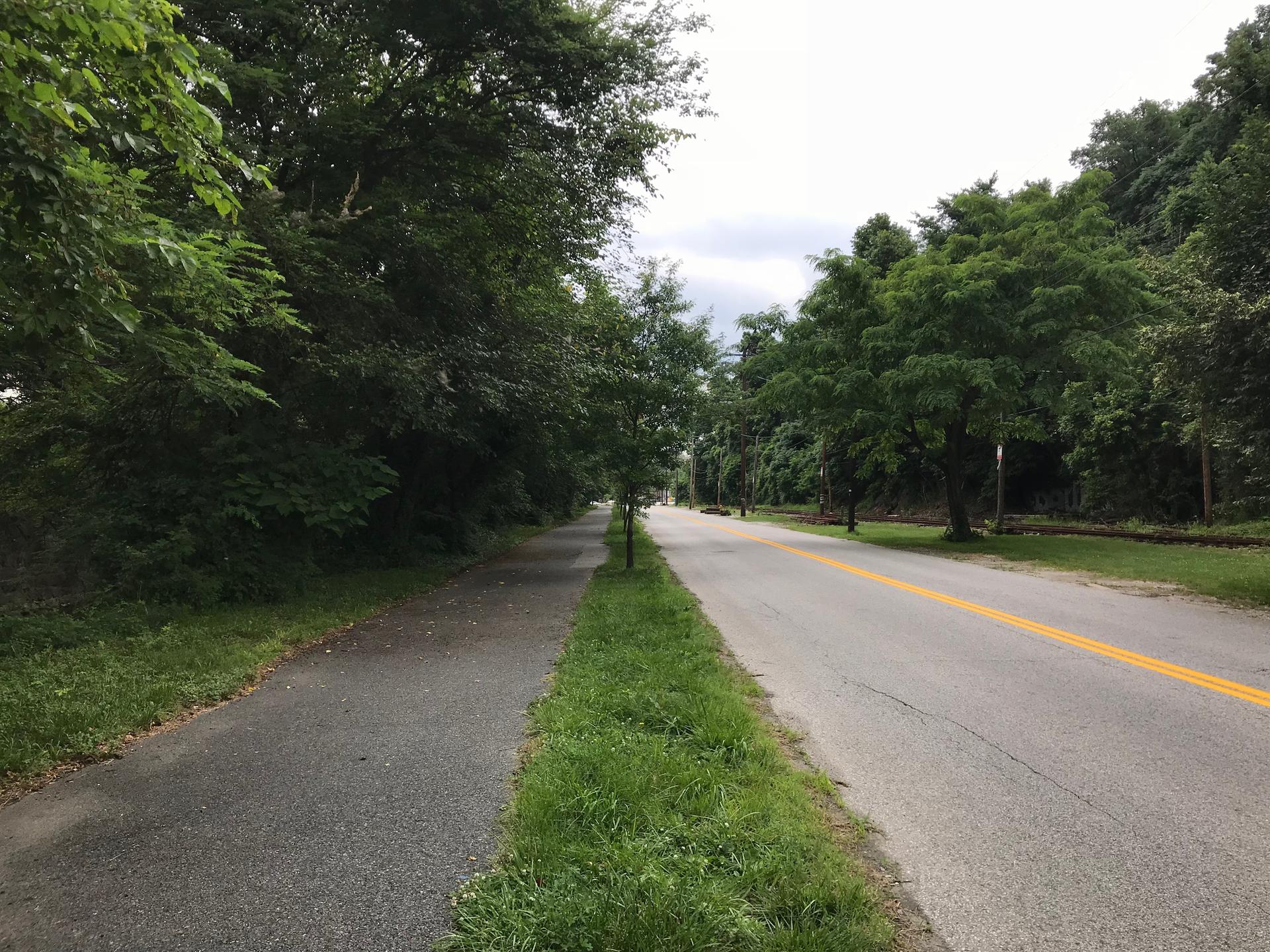 A paved road runs alongside a walking path, bordered by green grass and trees.