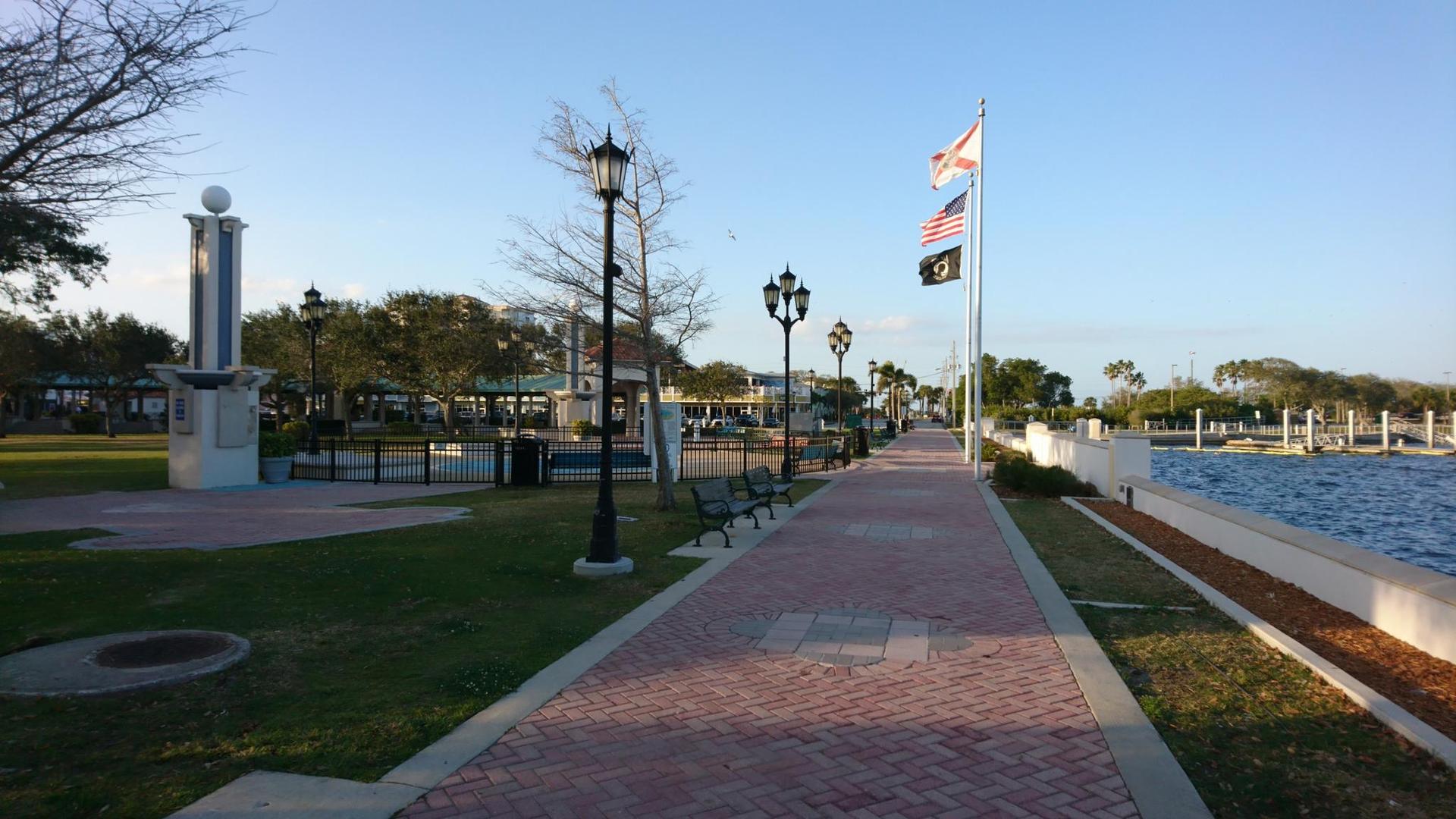Sunny paved promenade next to water with flags flying and park benches.