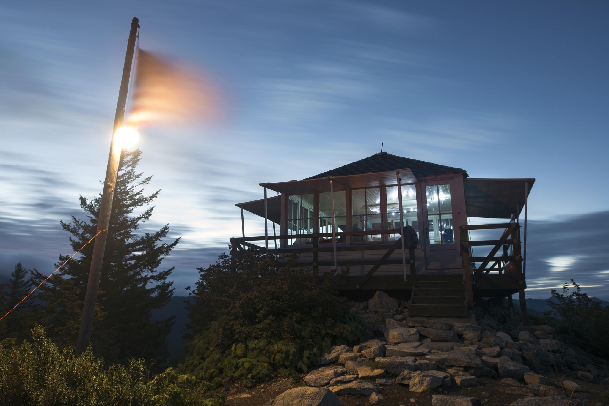 Wooden lookout tower and bright light pole at dusk on a rocky hilltop.