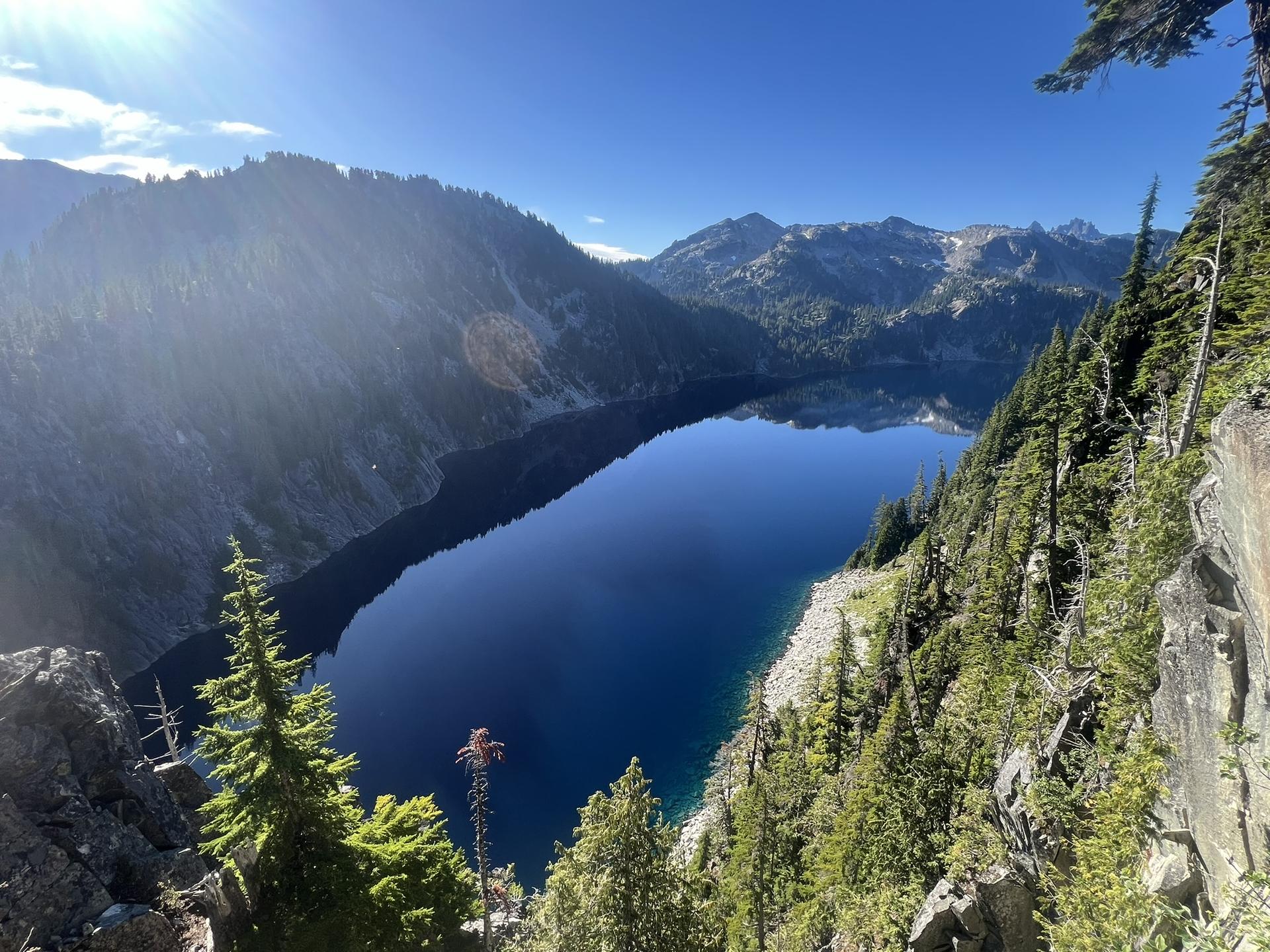 Deep blue mountain lake surrounded by steep, forested slopes under bright sunlight.