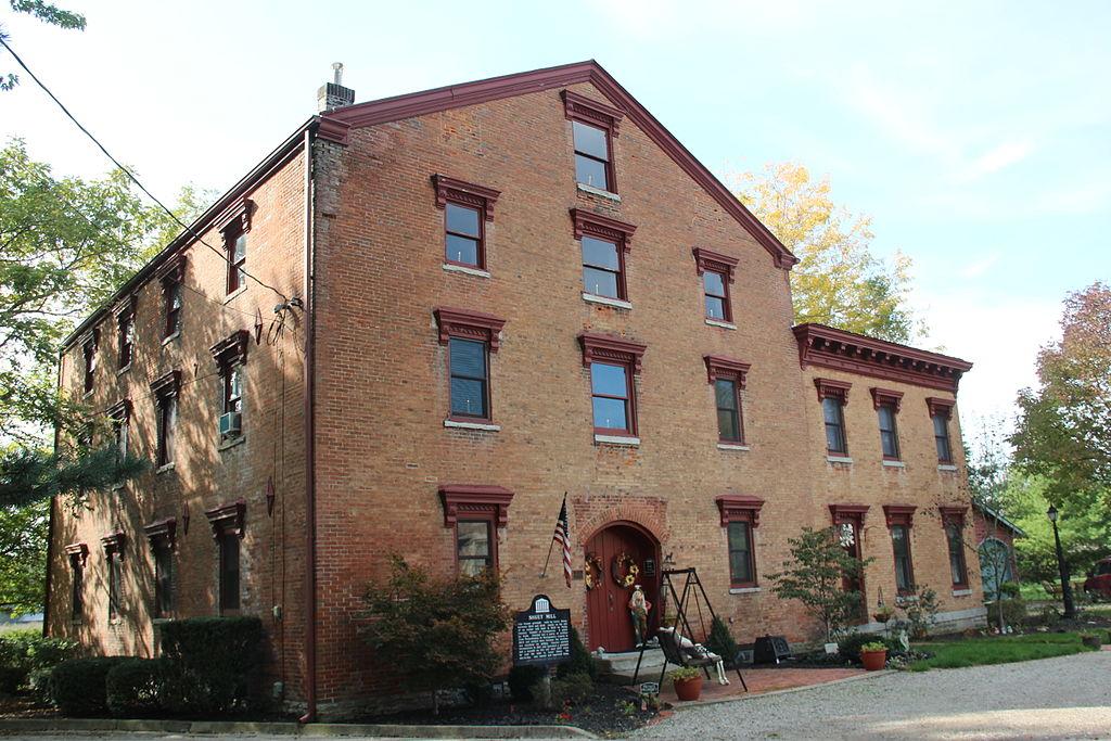 Historic brick building with many windows, surrounded by autumn trees in daylight.