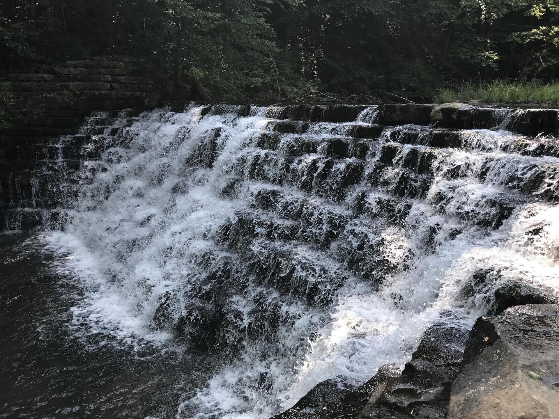 Waterfall cascades over stepped rocks with green trees in natural light.