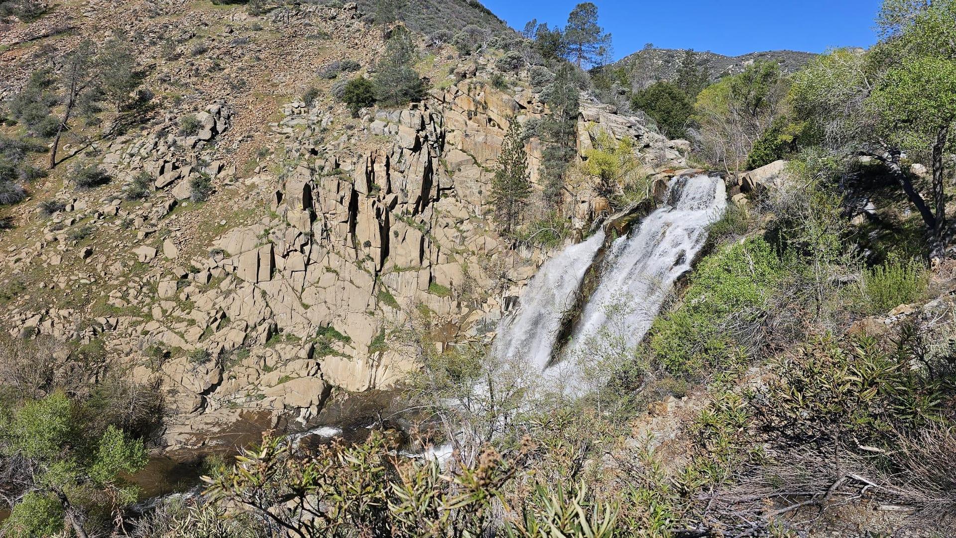 Waterfall cascading down a rocky cliff face, surrounded by green shrubs, sunny.