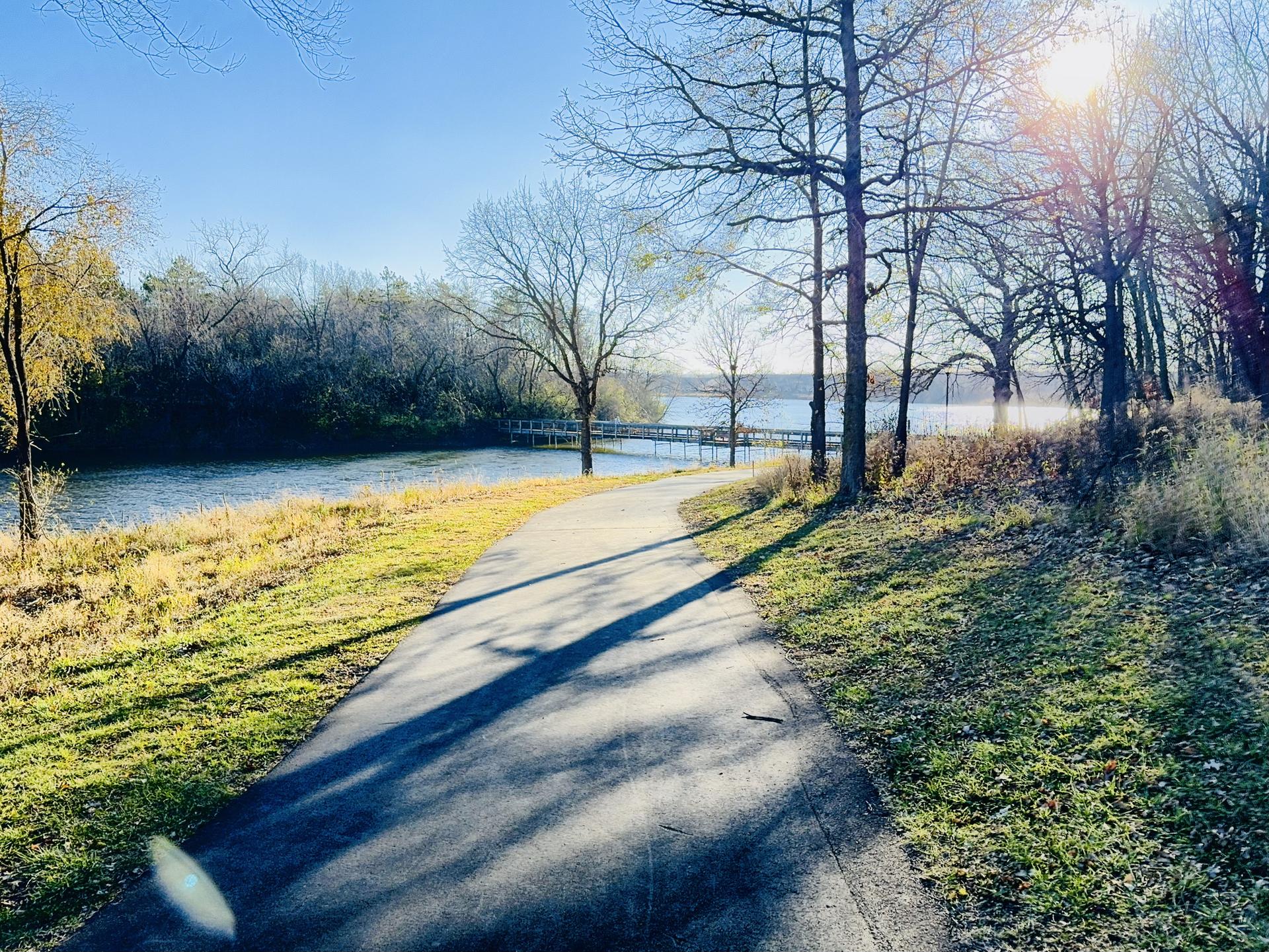 A sunny paved path alongside a lake with long tree shadows in autumn.