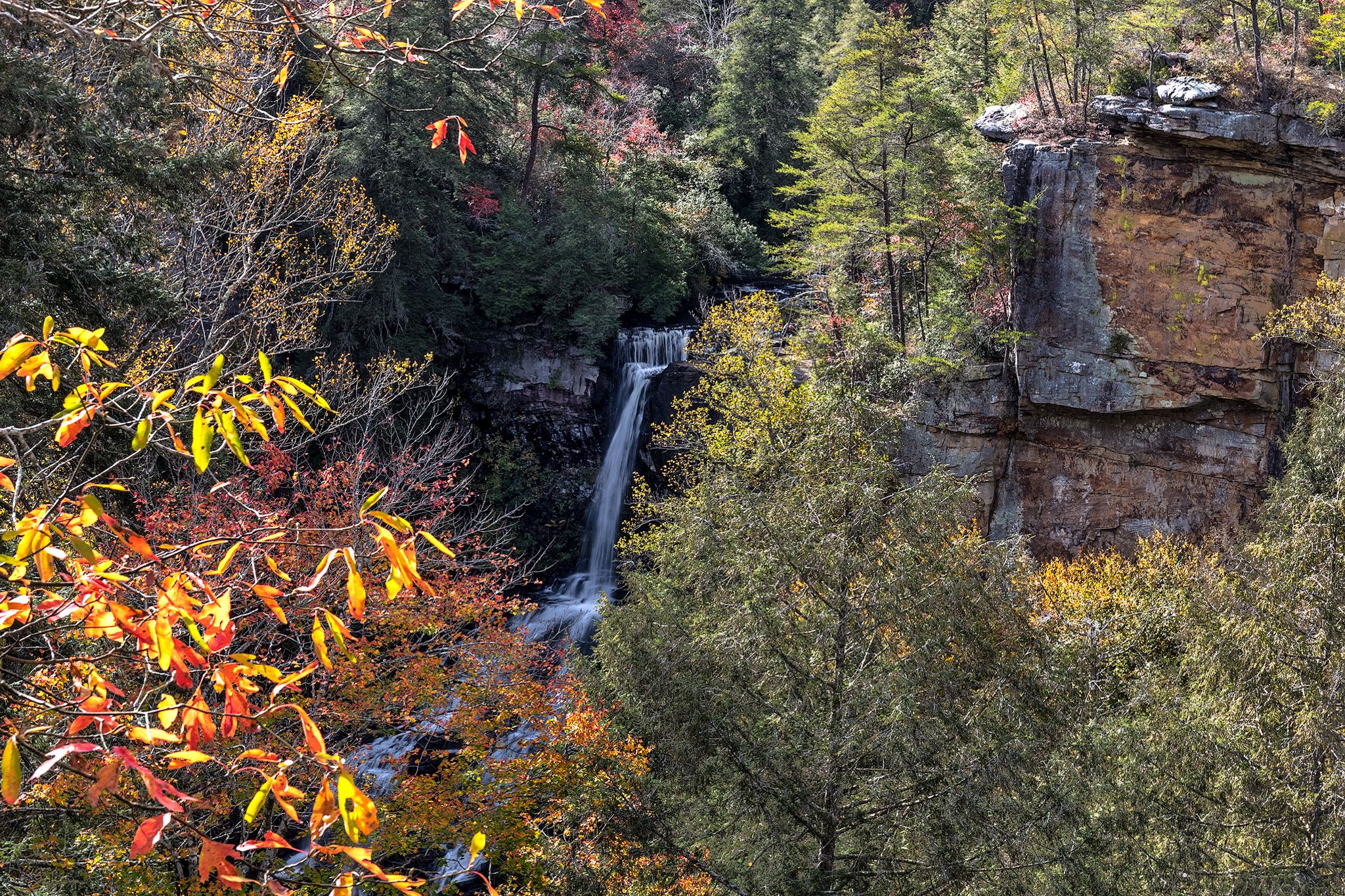 A waterfall cascades down rocky cliffs surrounded by vibrant autumn forest foliage. - Lower Loop Trail