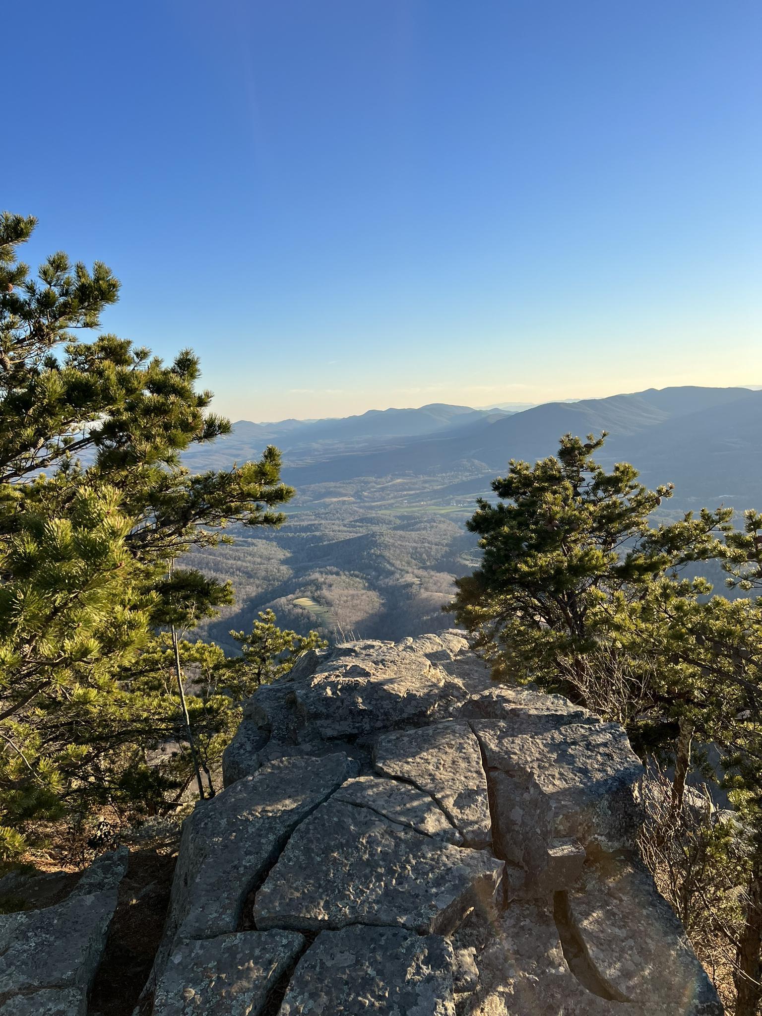Sunny mountain vista with rocky foreground and distant valleys under clear sky.