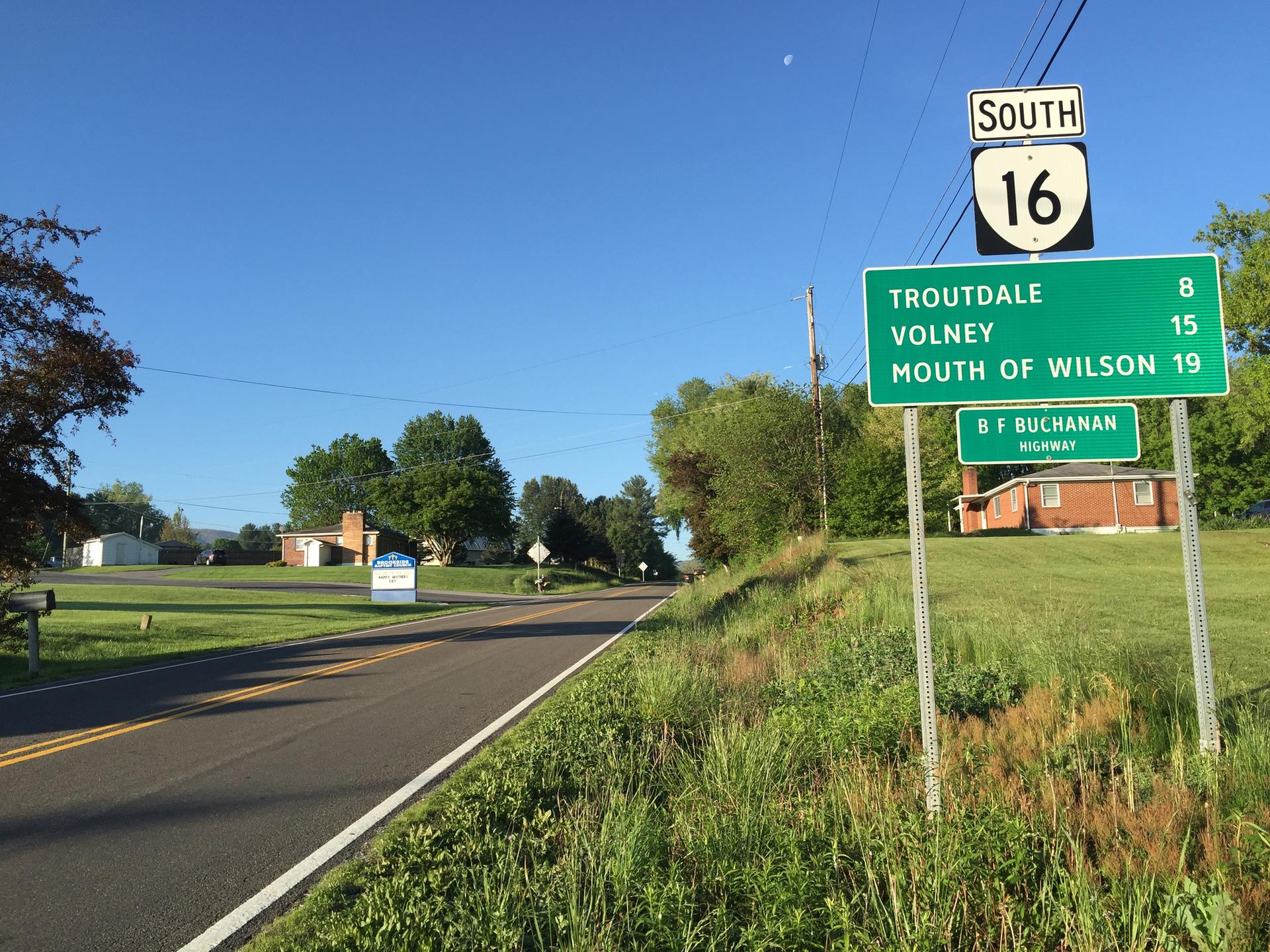 Road signs beside a paved road in a sunny, green rural landscape.