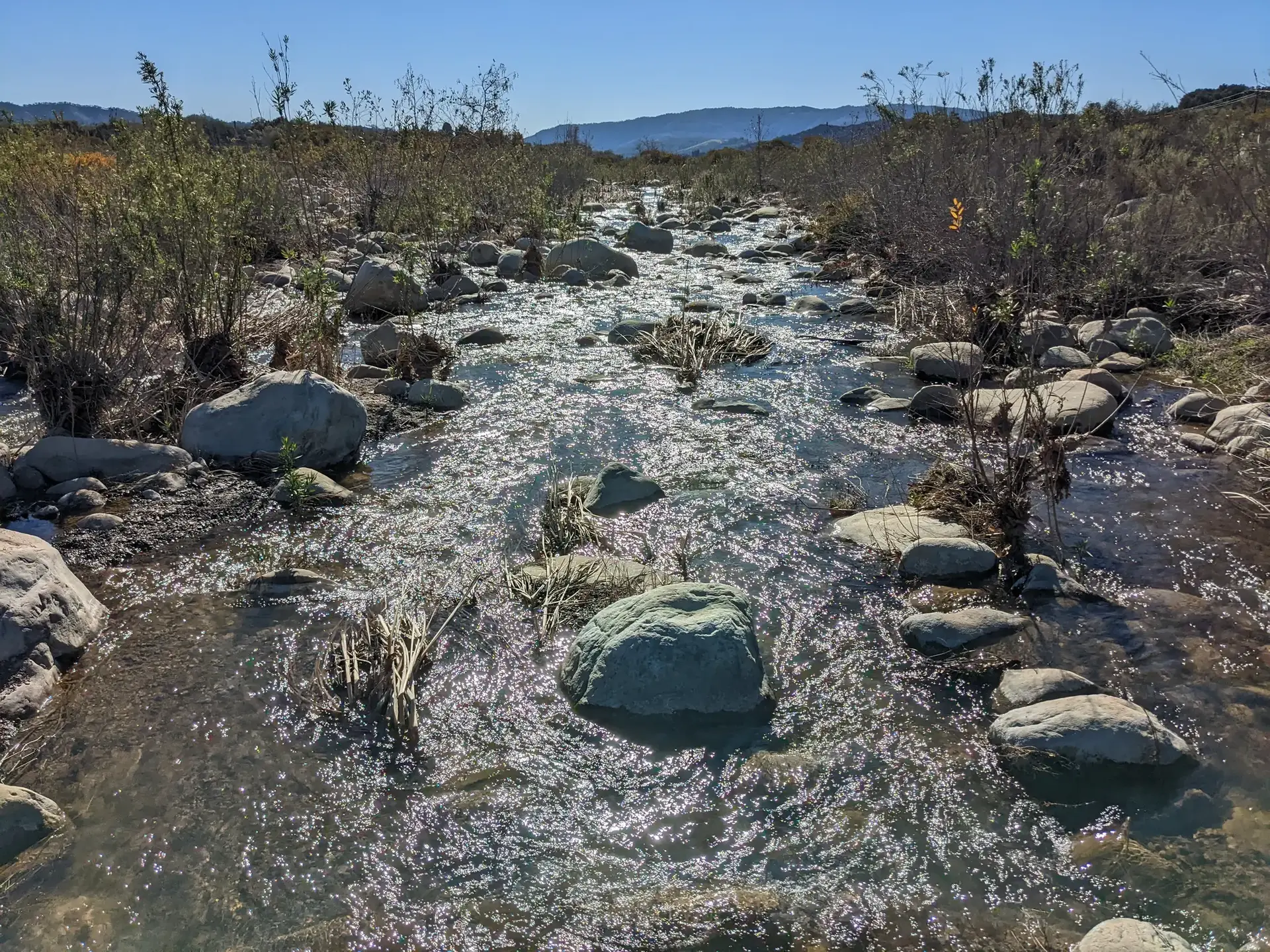 Bright sunlight sparkles on a rocky stream flowing through an arid landscape.