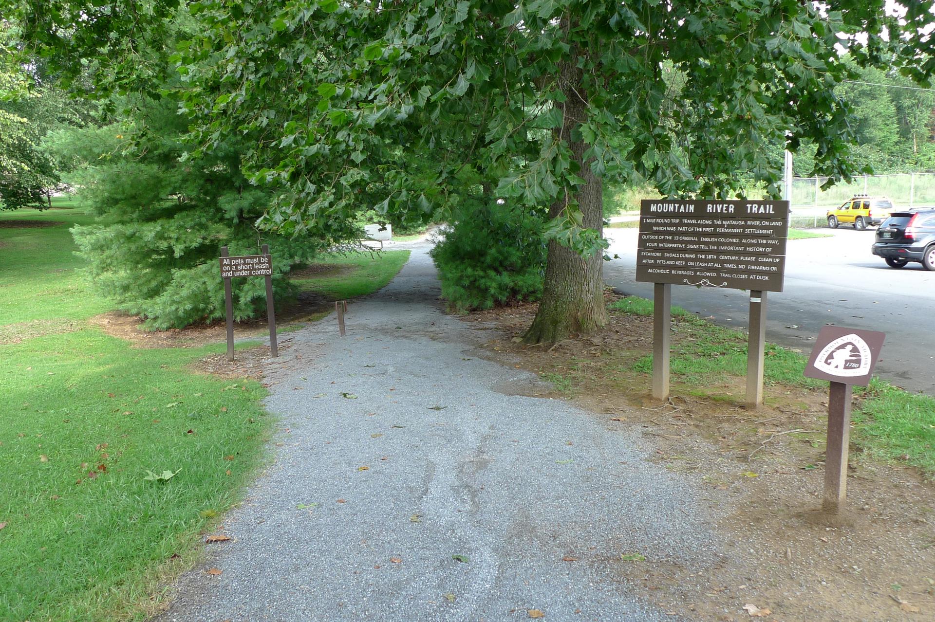 Gravel path leading into shaded woods with informational signs and distant cars.