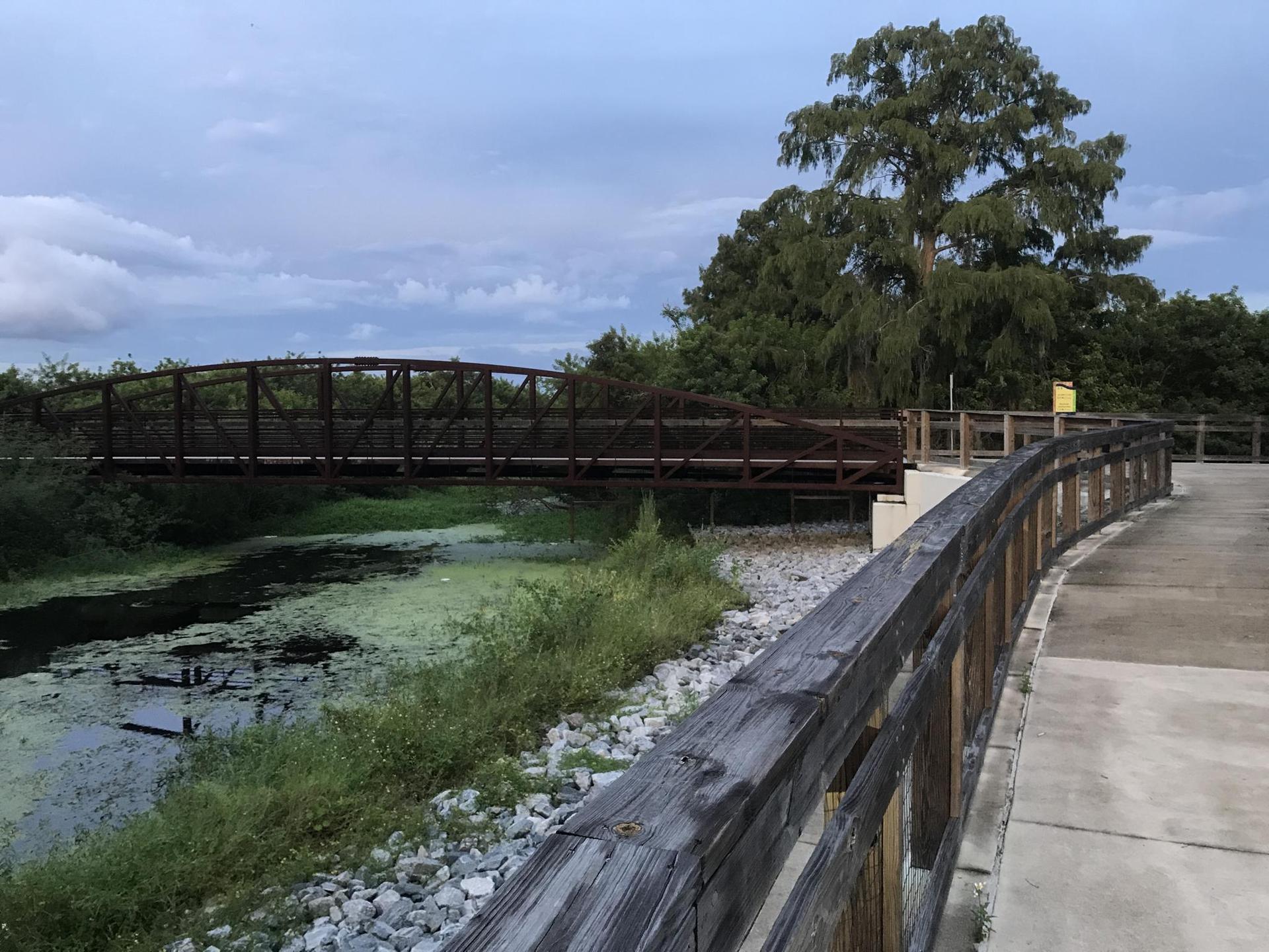 Wooden boardwalk leading to a metal bridge over murky water at dusk.