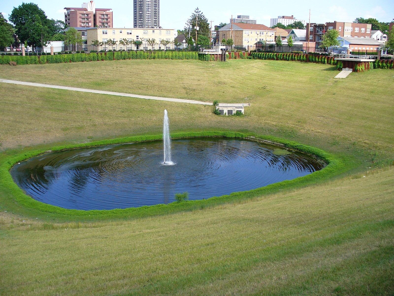 A small pond with a fountain in a large green park with distant buildings.